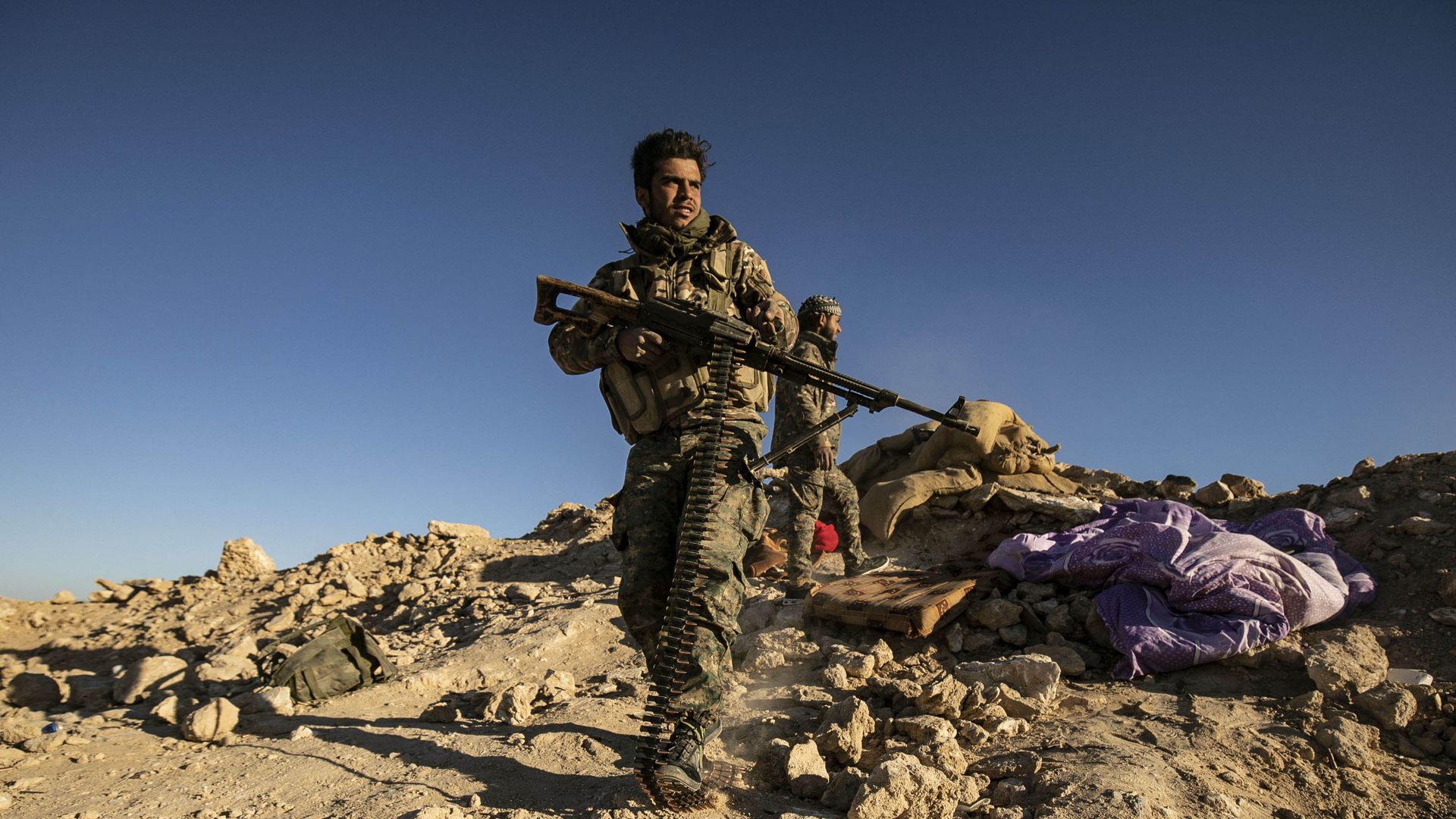 A Syrian fighter stands alone on a dirt hill, carrying an assault weapon.