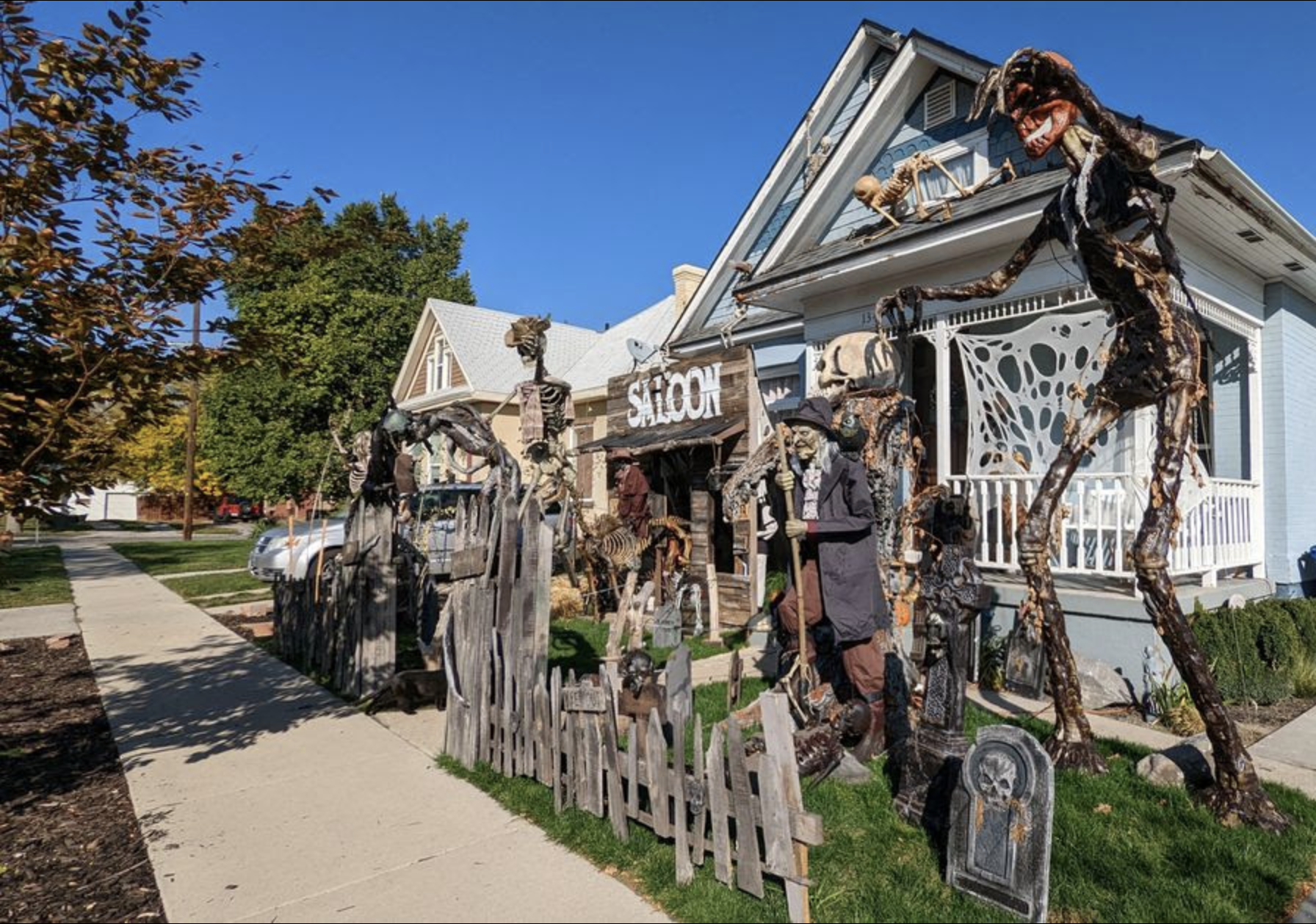 Wide shot of a blue house covered in Halloween decorations: a towering monster on the roof, skeletal figures along a wooden fence, and a rustic Saloon sign in the yard.