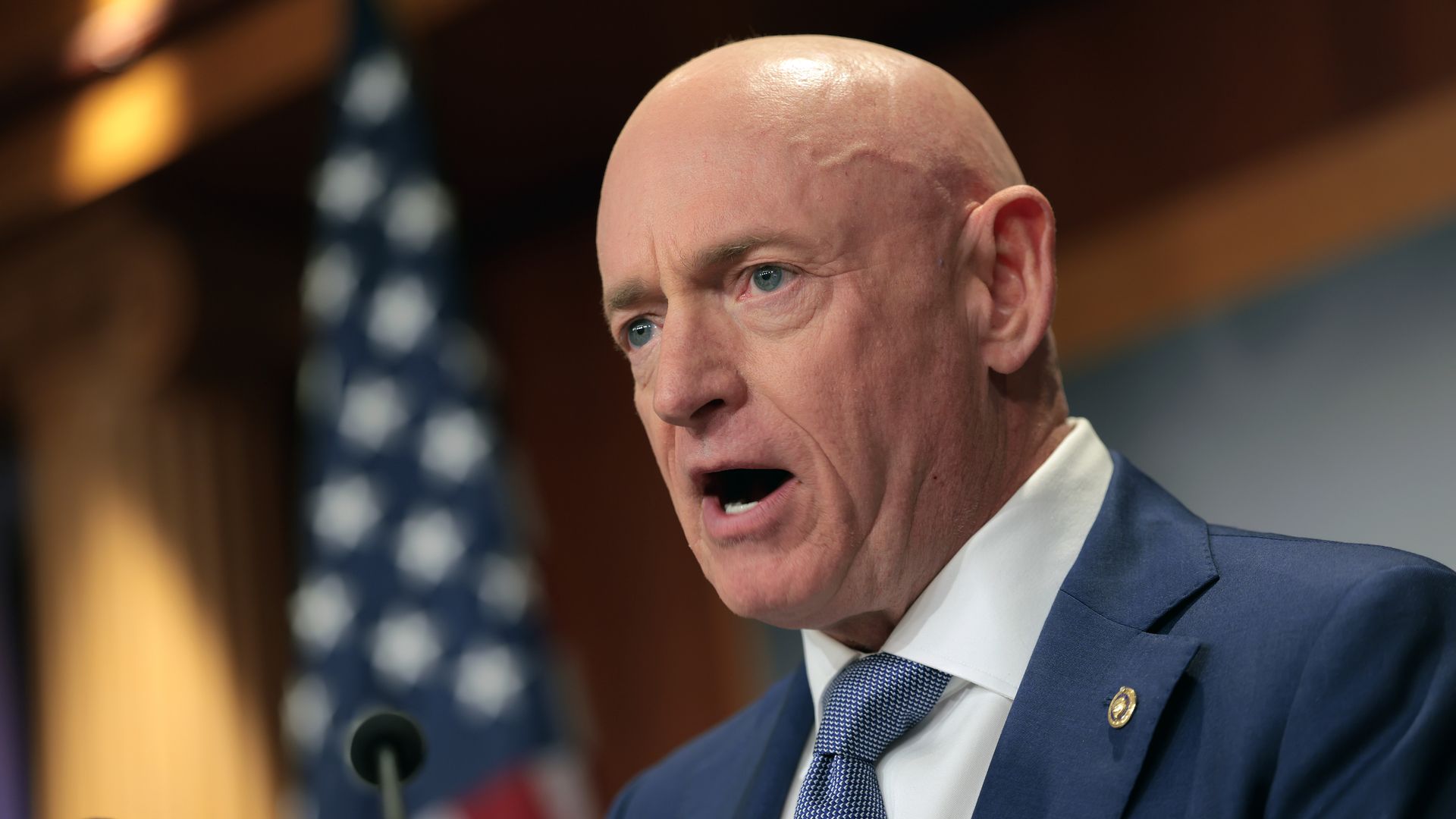 A bald man in a navy blue suit jacket with a pin on the lapel and lighter blue tie over a white shirt opens his mouth as he speaks into a microphone, with a United States flag in the background.