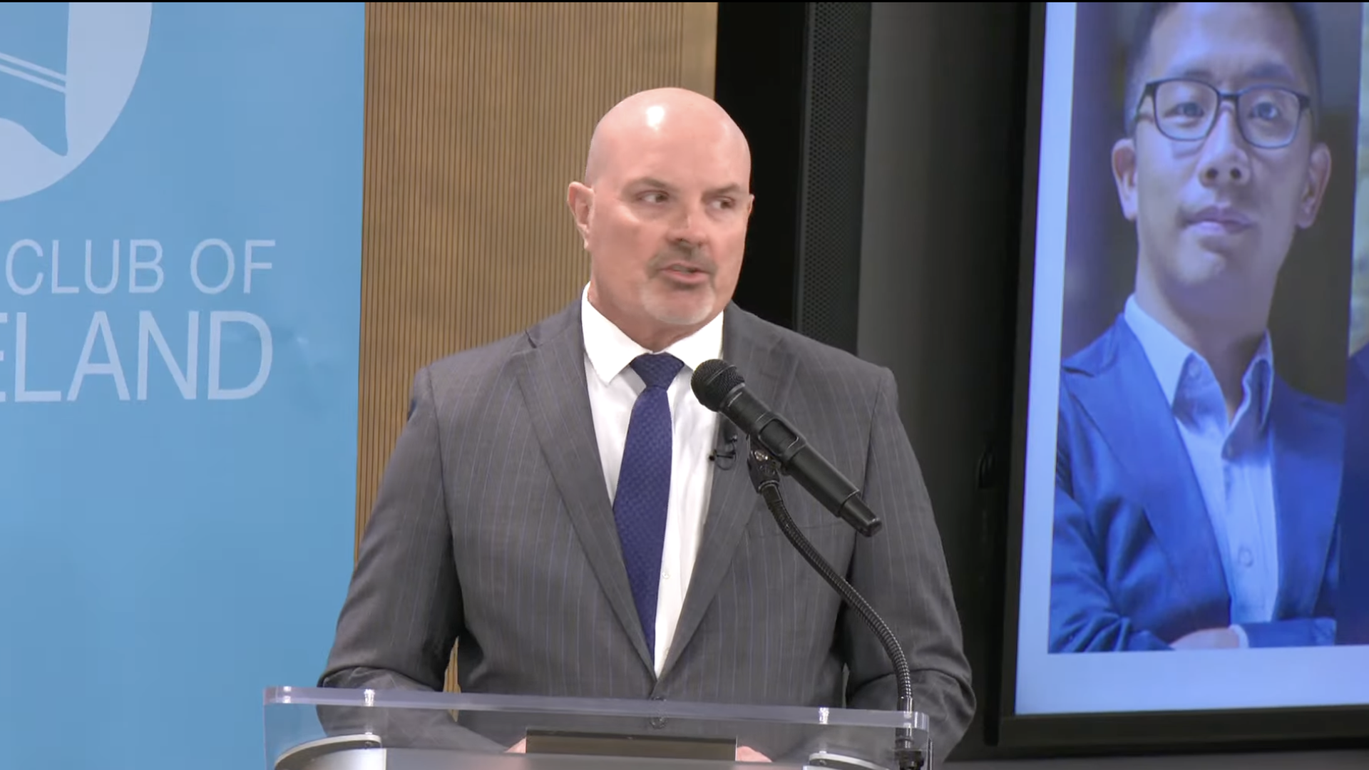 A screenshot of a suited prosecutor speaking at a lectern wih the image of his challenger over his shoulder