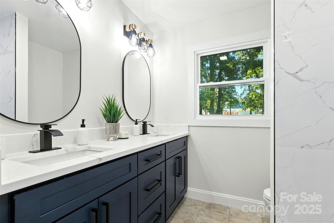 Bright modern bathroom with dark blue double vanity, white marble countertop, two black-framed oval mirrors, black faucets, pendant lights, green plant, window with tree view.