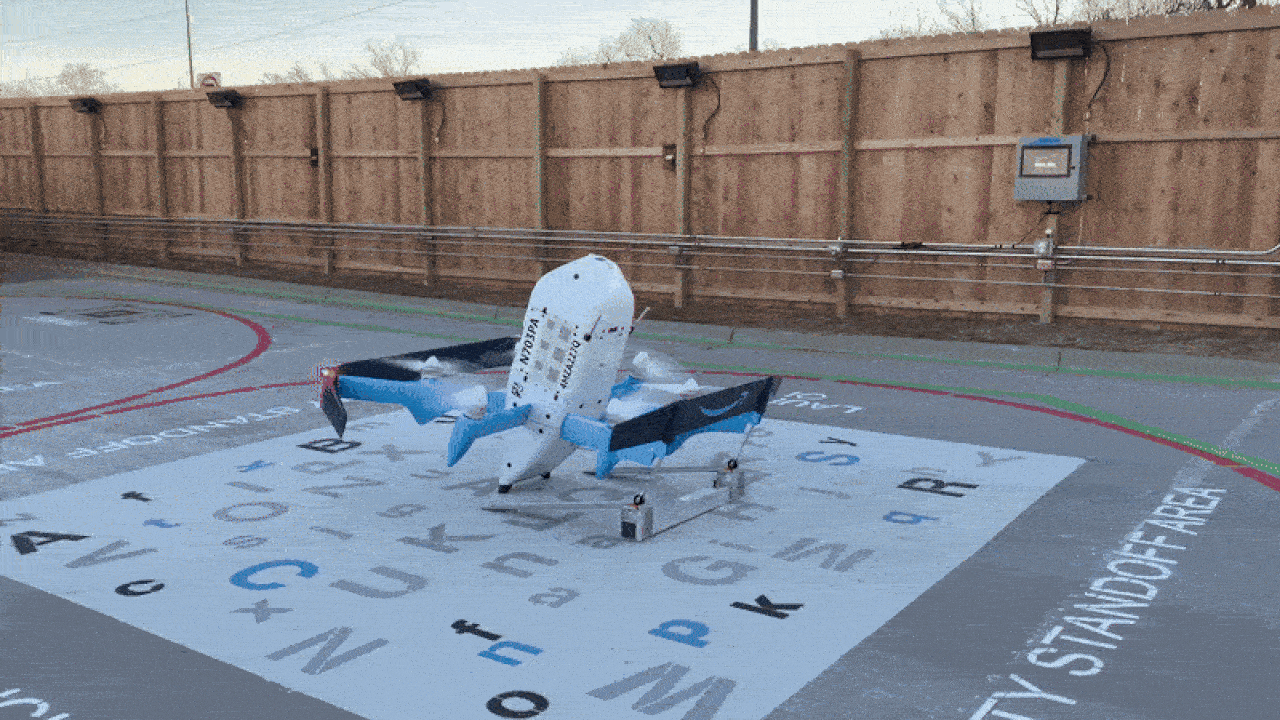 A white and blue drone with multiple propellers stands on a landing pad marked with various letters and labeled "SAFETY STANDOFF AREA" on the right, surrounded by a wooden fence.