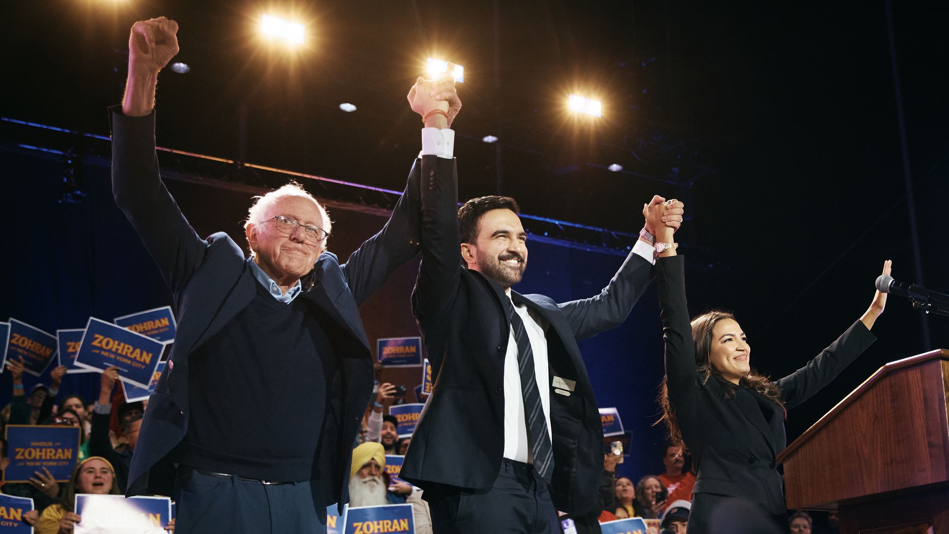 Bernie Sanders, Zohran Mamdani and Alexandria Ocasio-Cortez raise their arms together in front of a crowd.