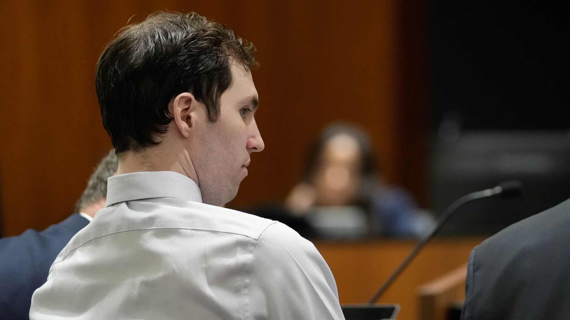 Side profile of a man in a light gray shirt seated in a courtroom. Warm brown wood-panel backdrop, with a dark area to the right, and a microphone in the foreground.
