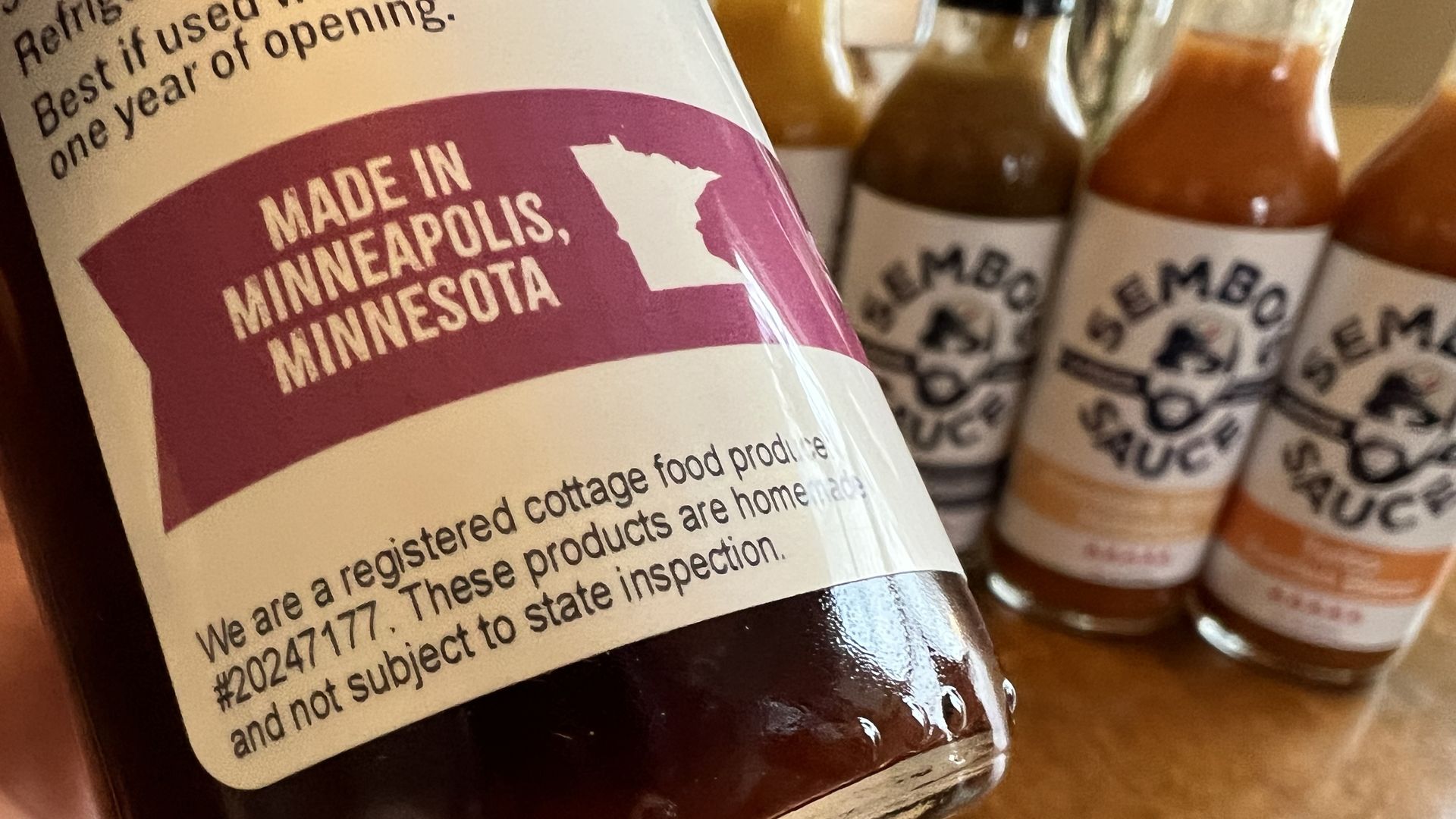 The label of a bottle of hot sauce held close to the camera. The text reads 'Made in Minneapolis, Minnesota; we are a registered cottage food producer.' Other bottles of hot sauce are arranged in the background.