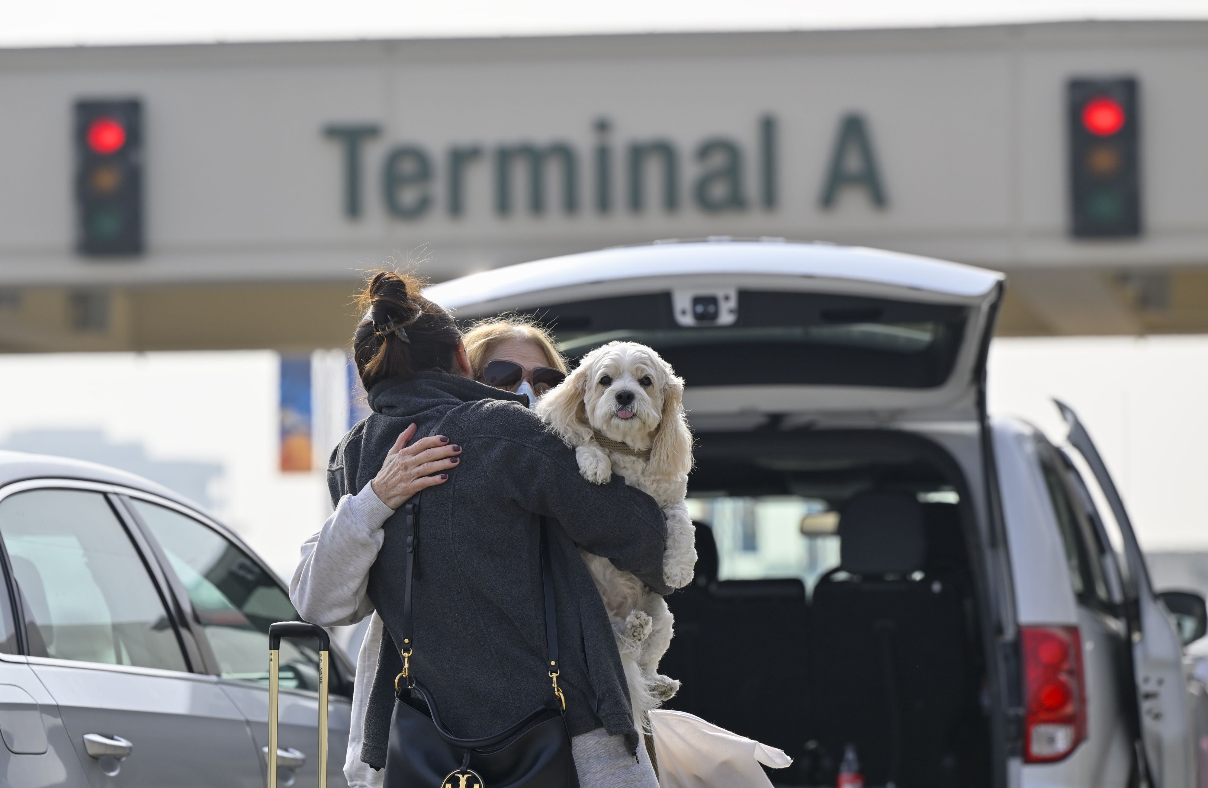 Photo of two people hugging with one holding a dog that peers over their shoulder, outside an airport terminal