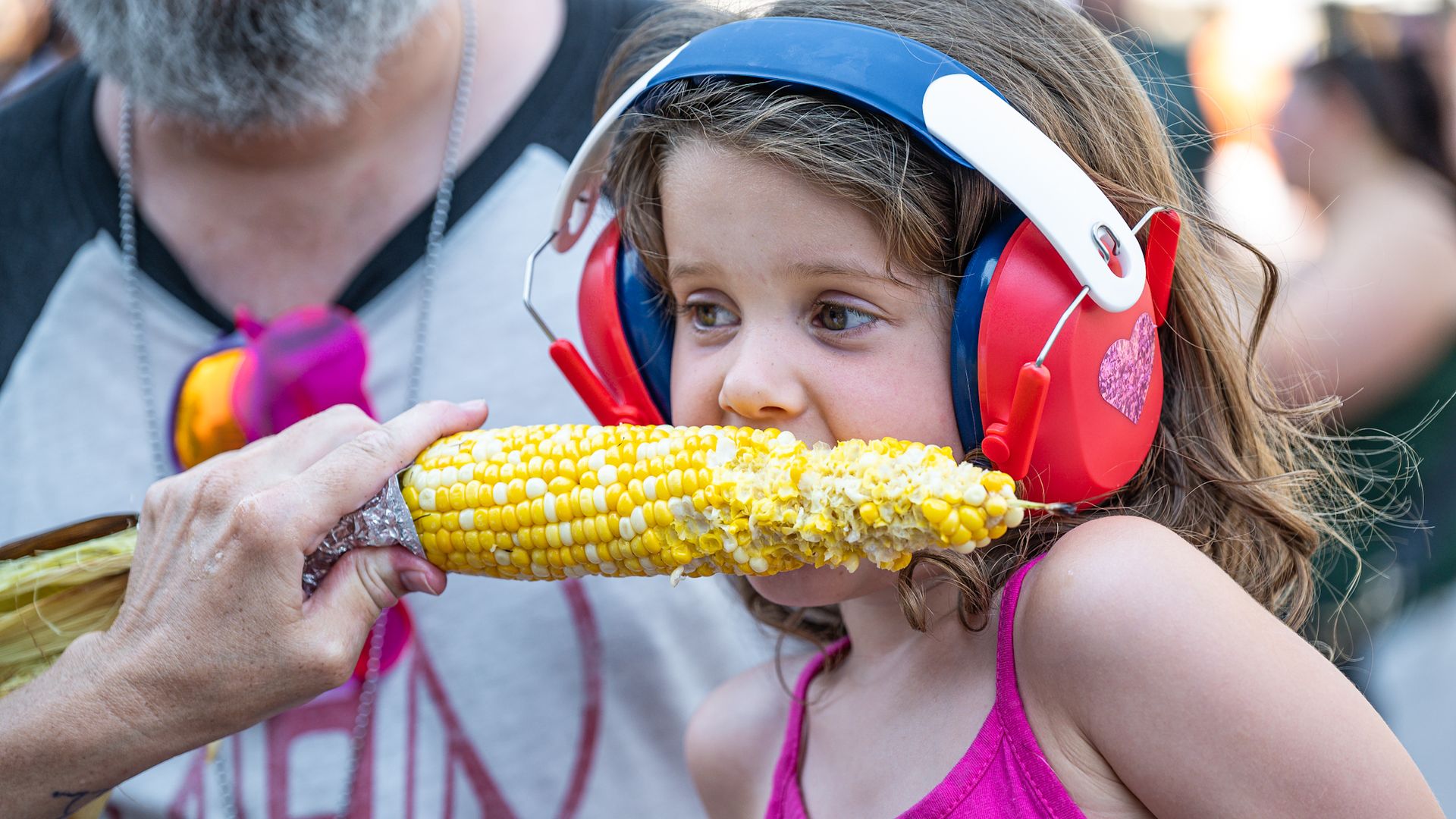 Young girl in a pink tank top wears blue and red earmuffs while biting into a corn cob handed to her by an adult in a gray shirt. Bright yellow kernels, outdoor scene.