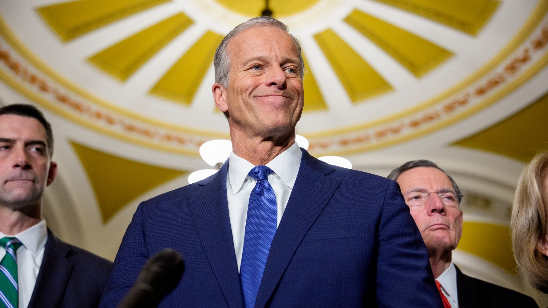 Smiling older man in a blue suit and blue tie stands at a podium, flanked by two men in suits, with a gold sunburst ceiling in the background.