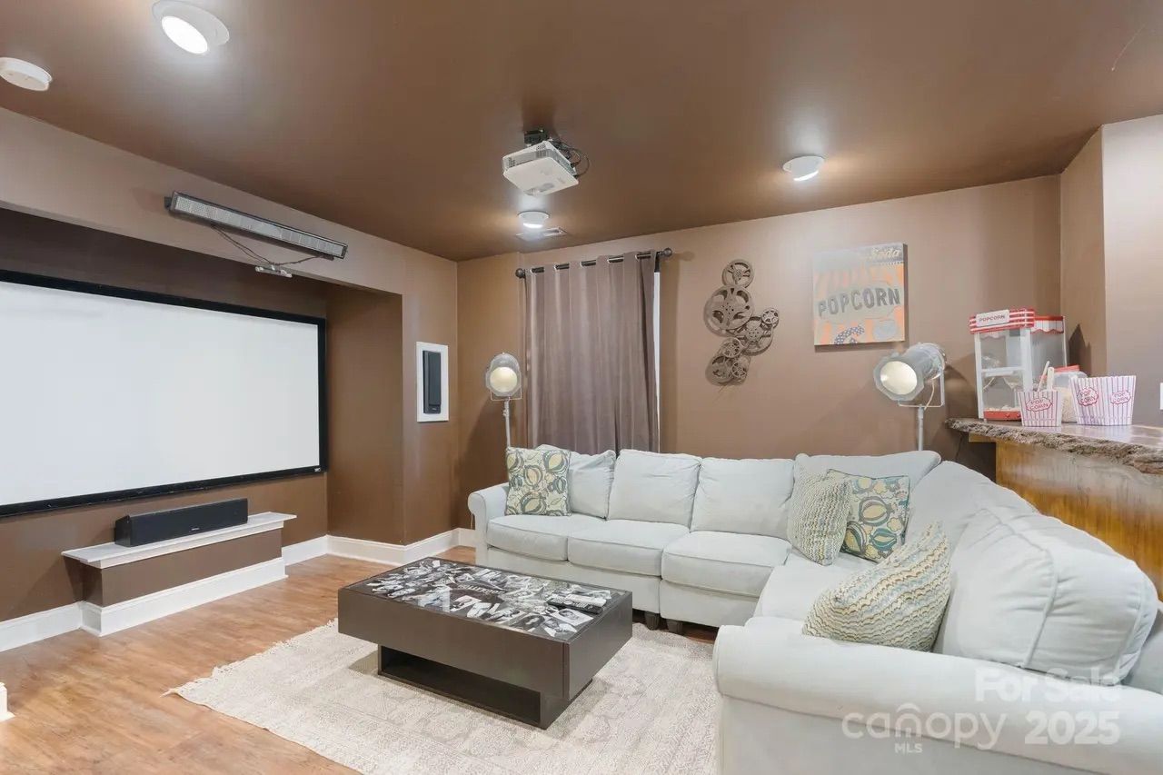 Cozy home theater room with brown walls and ceiling, large white sectional sofa with patterned pillows, black coffee table, projector screen, floor lamps, and popcorn machine on counter.