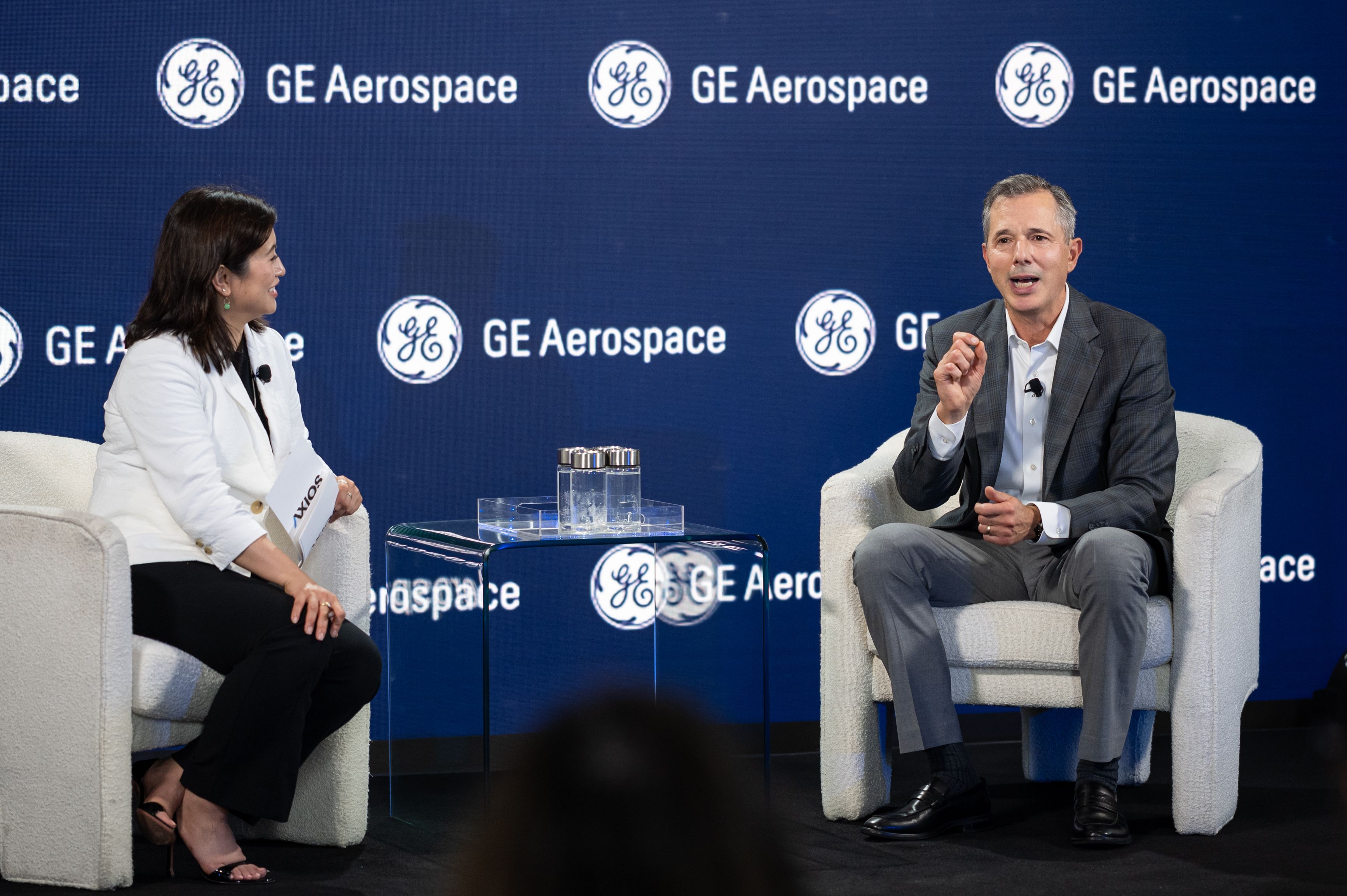 Two people sit on stage in cream chairs during a panel, a woman in a white blazer on left and a man in a gray suit on right; blue backdrop with GE Aerospace logos, glass coffee table between them.