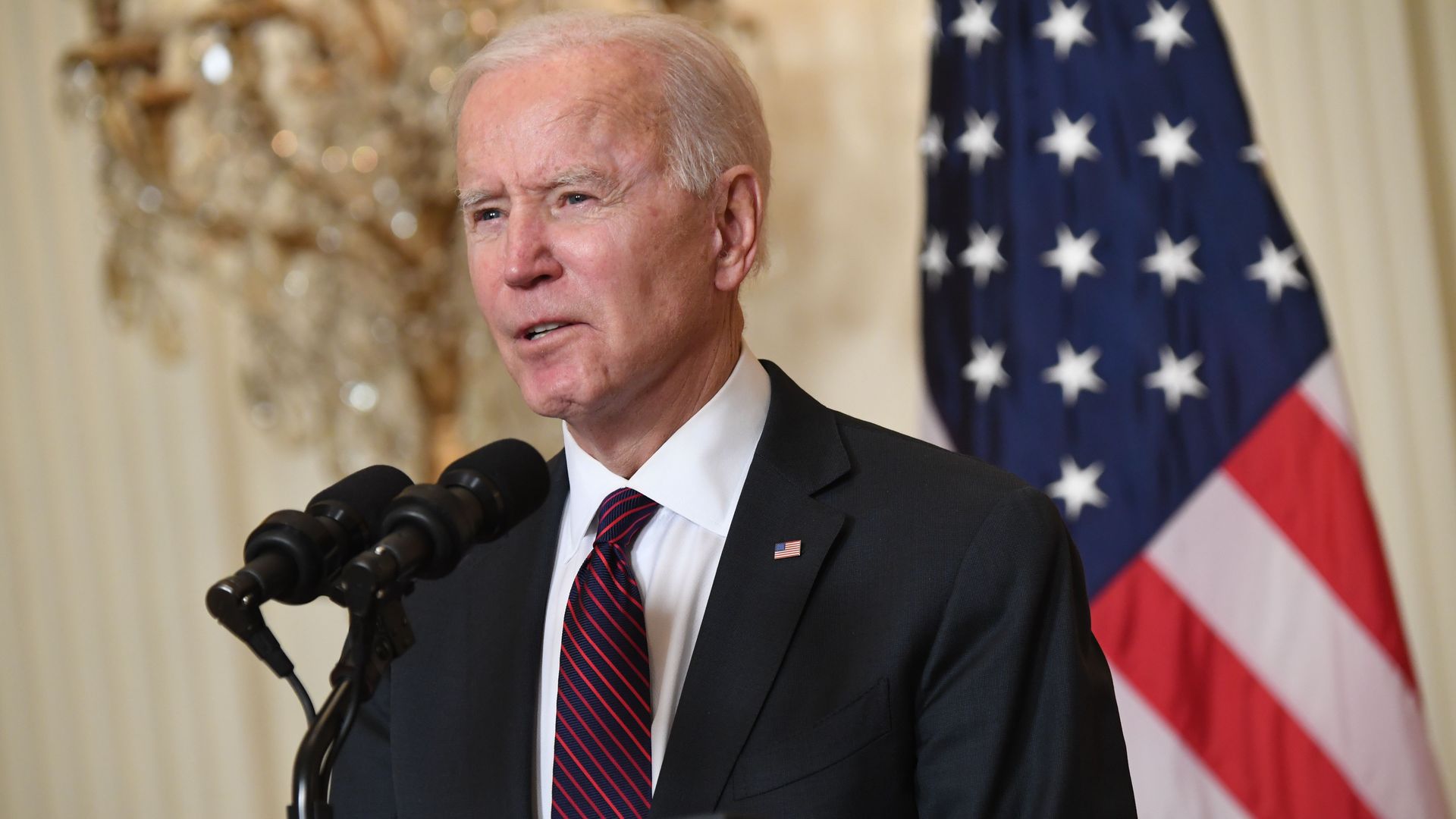 Biden wears a suit and stands in front of an American flag