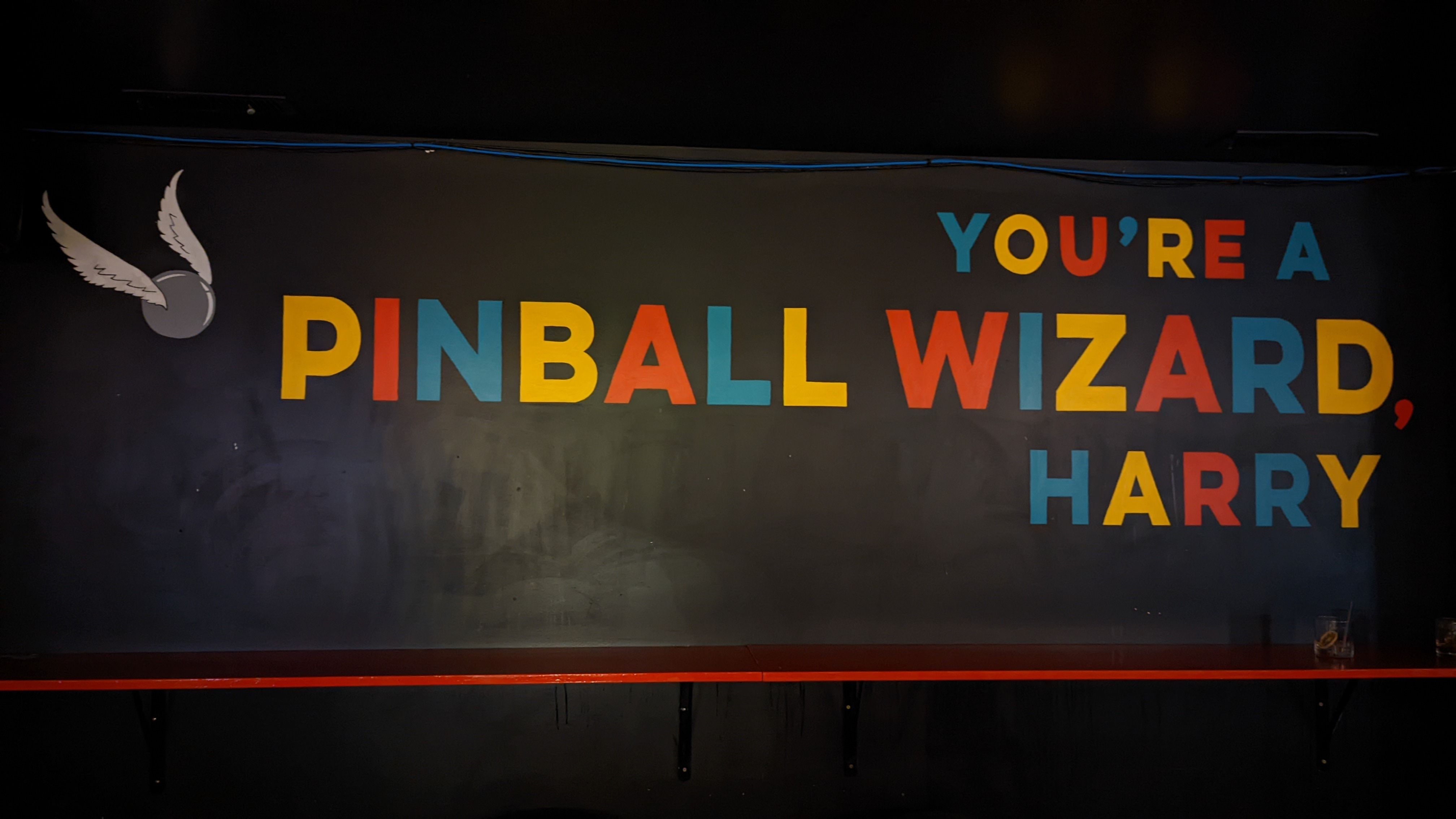 A sign says "You're a pinball wizard, Harry" in multi-colored letters, behind a shelf with a cocktail on it.