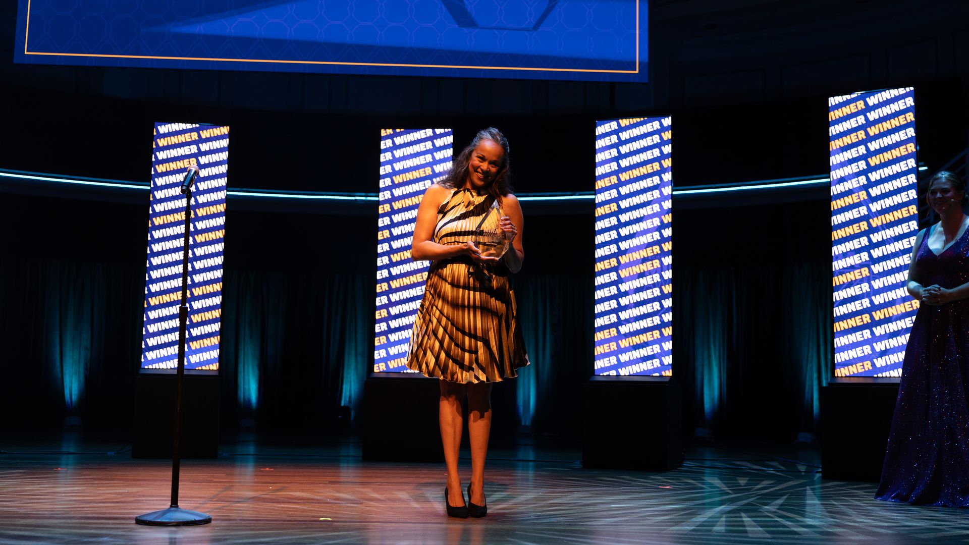Smiling woman in a striped dress stands center stage, holding an award trophy. Blue-lit stage with "WINNER" panels behind her and a microphone stand in front.