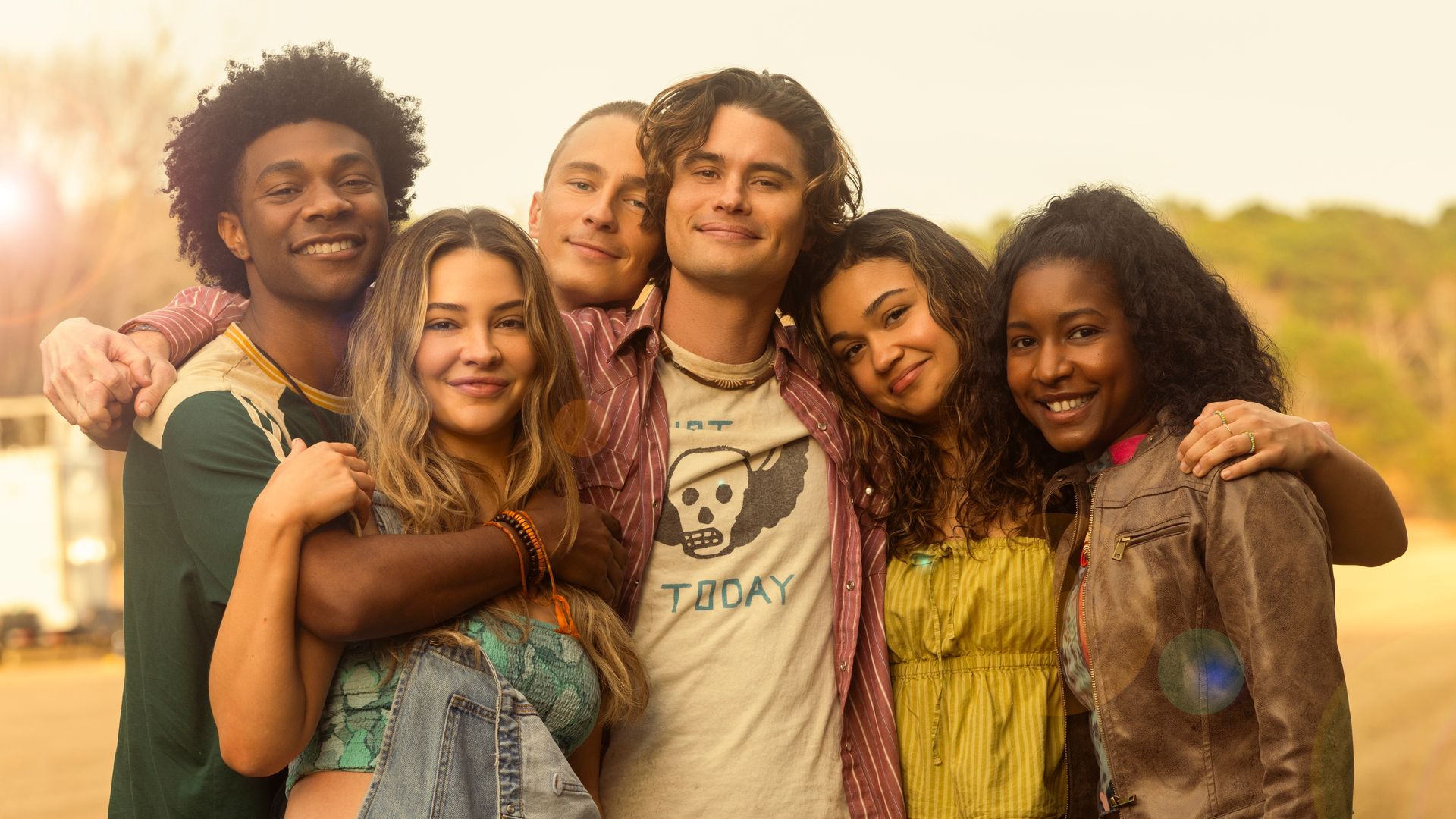 Six young people — played by Jonathan Daviss, Madelyn Cline, Drew Starkey, Chase Stokes, Madison Bailey and Carlacia Grant — stand close together outdoors, smiling with arms around each other on a sunny day.