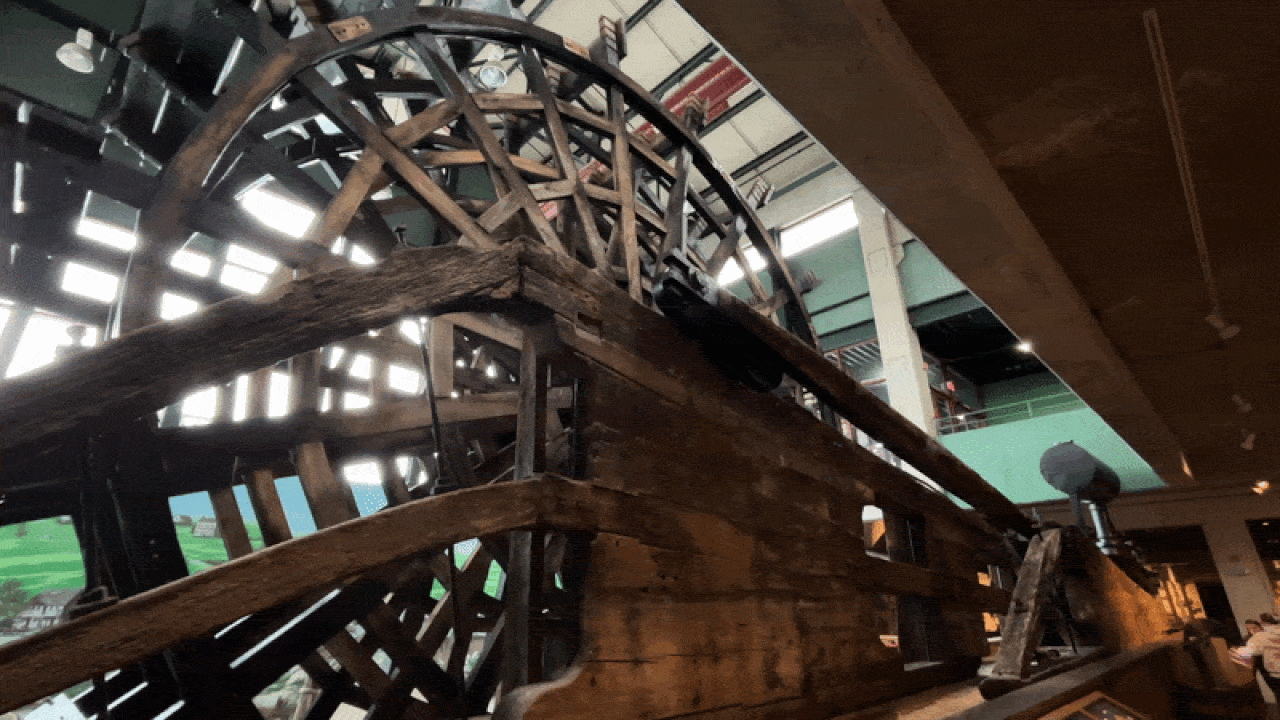 Inside a museum, a massive weathered wooden water wheel with lattice spokes dominates the scene, supported by thick timber beams, with a modern interior visible in the background.