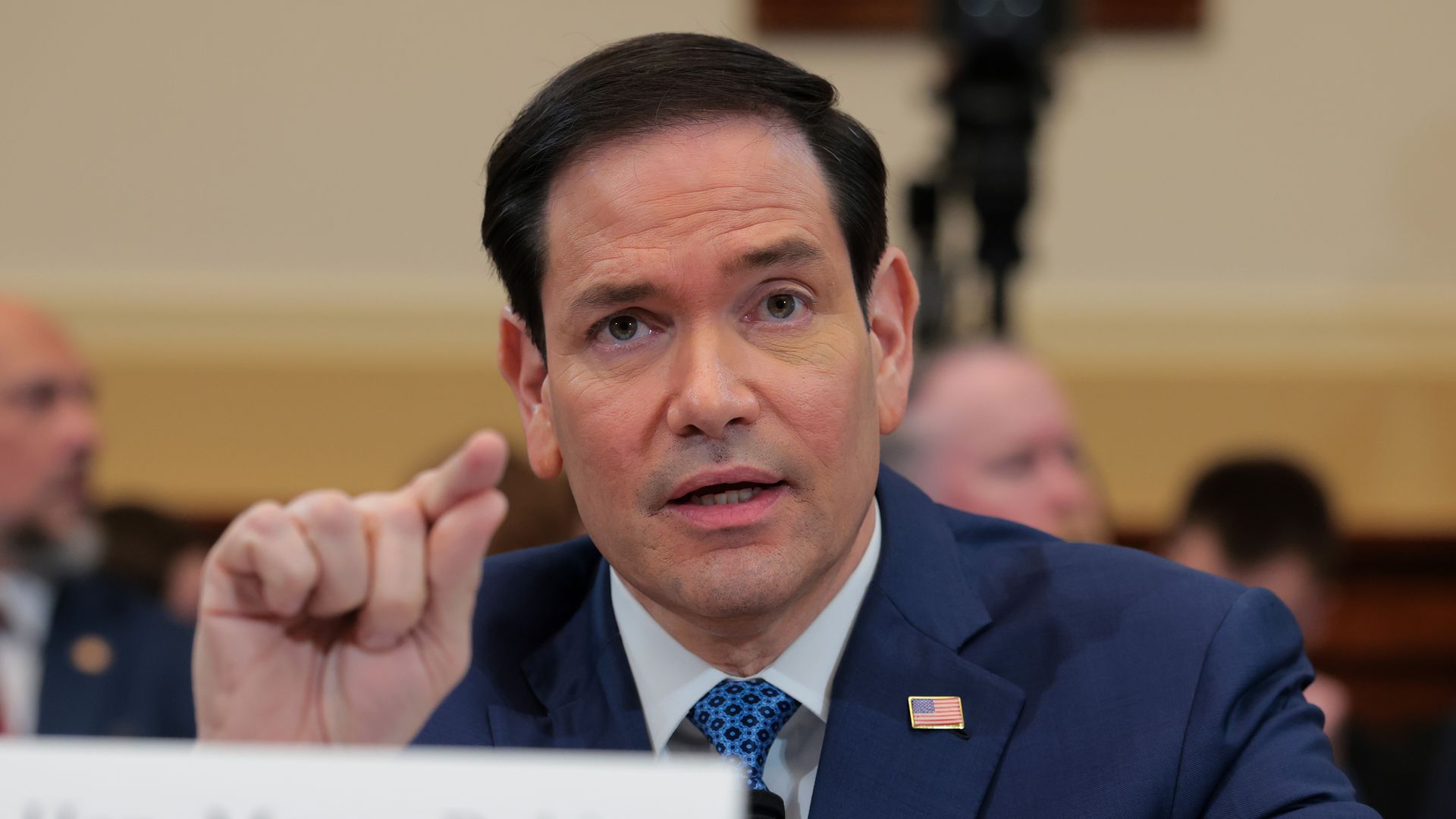 Secretary of State Marco Rubio — wearing a navy jacket, a white collared shirt, a blue tie and an American flag pin — points as he testifies to a House committee.