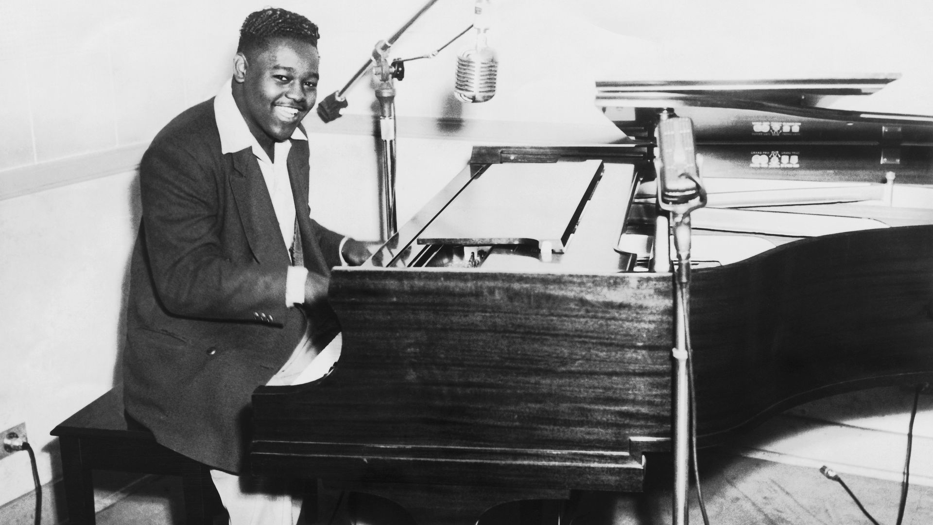 In a black and white photo, Fats Domino is seen playing piano in a recording studio while singing into a microphone.