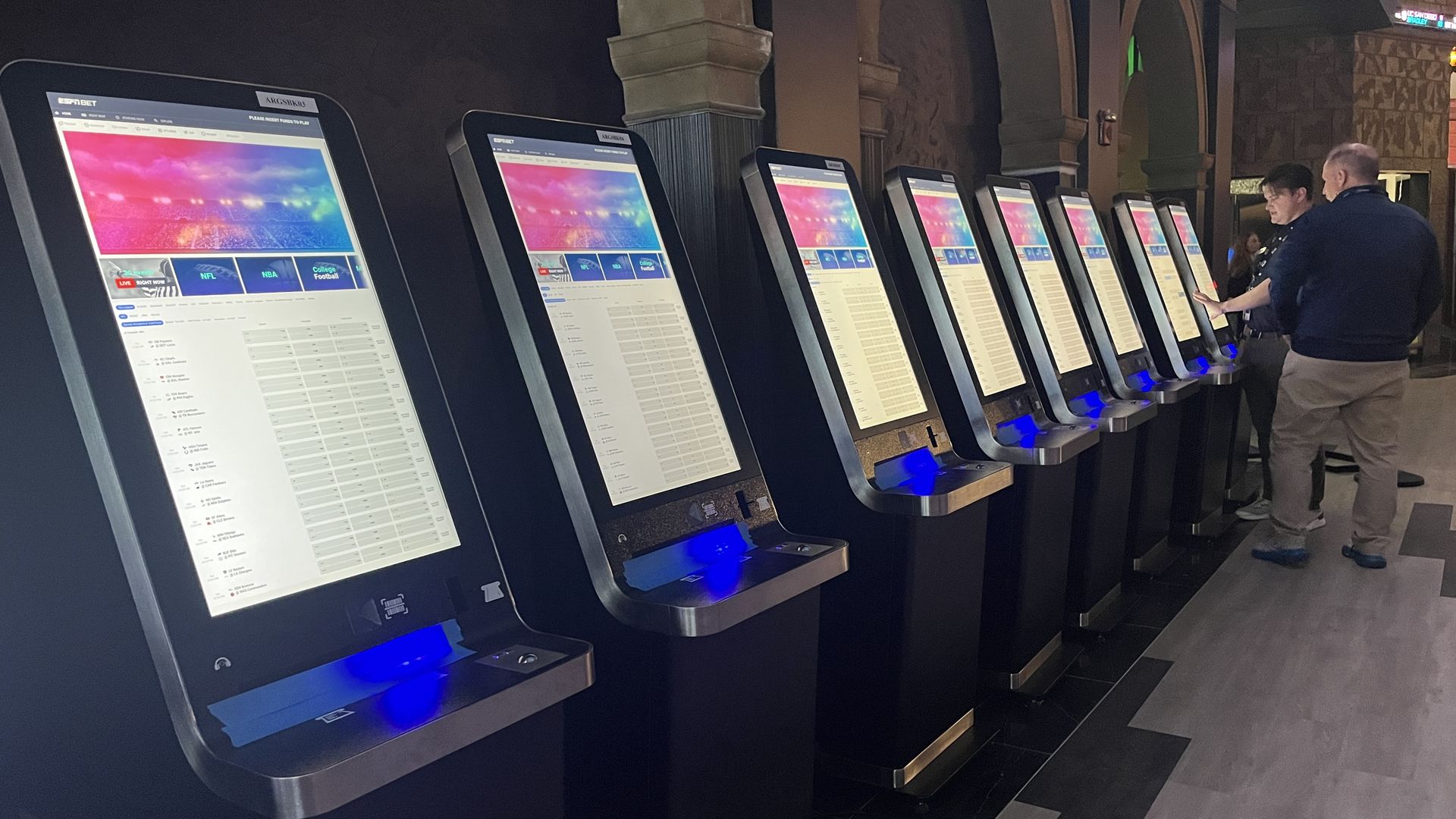 Row of black and silver touchscreen betting kiosks with colorful sports screens, two people interacting with one machine inside Argosy Casino.