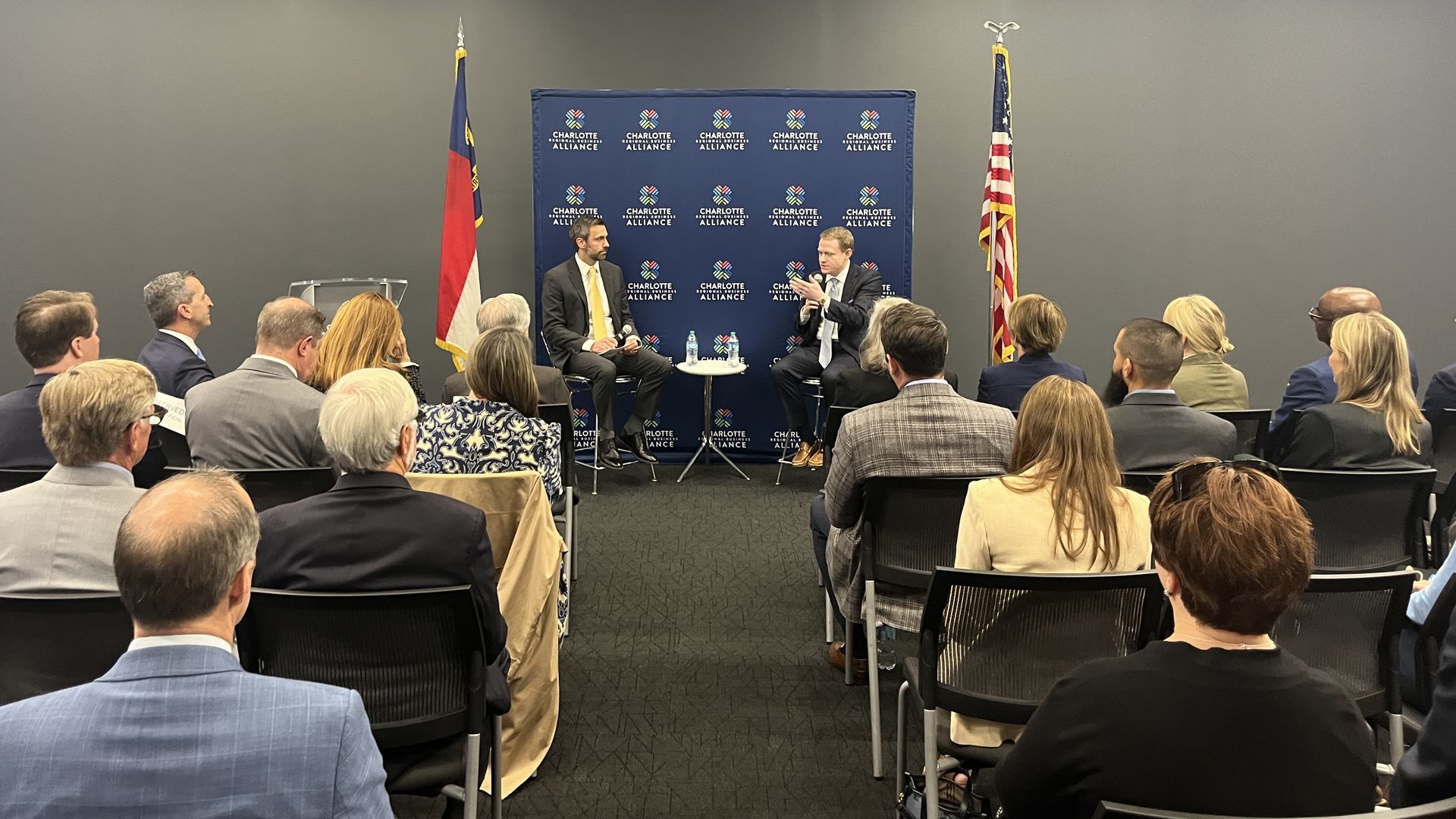 Two men in suits on a stage discuss with a blue Charlotte Alliance backdrop, flanked by a North Carolina flag and an American flag, as an audience watches a panel.