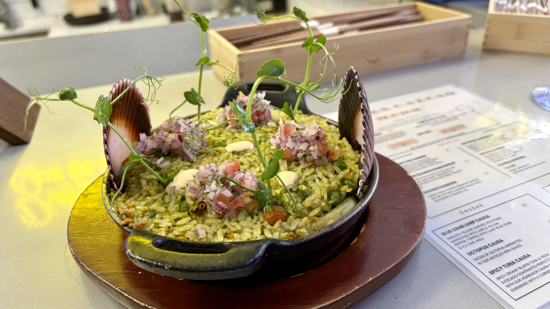 A shell-shaped bowl of rice topped with diced onions and herbs, finished with delicate pea shoots; a blurred kitchen counter and menu in the background.