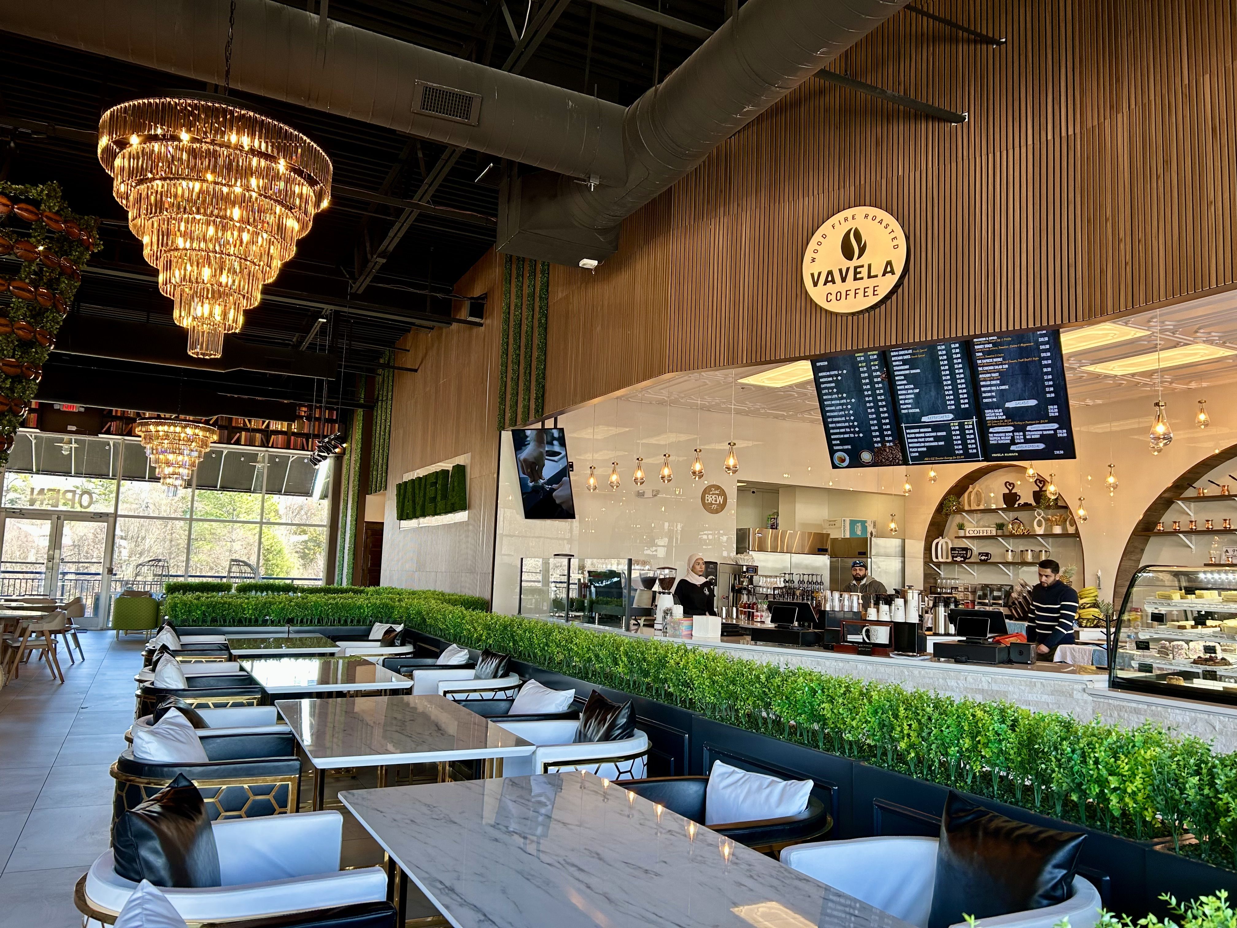 Modern coffee shop interior with marble tables, black and white chairs, green plants divider, large chandeliers, wooden wall with Vavela Coffee logo, and staff behind counter.
