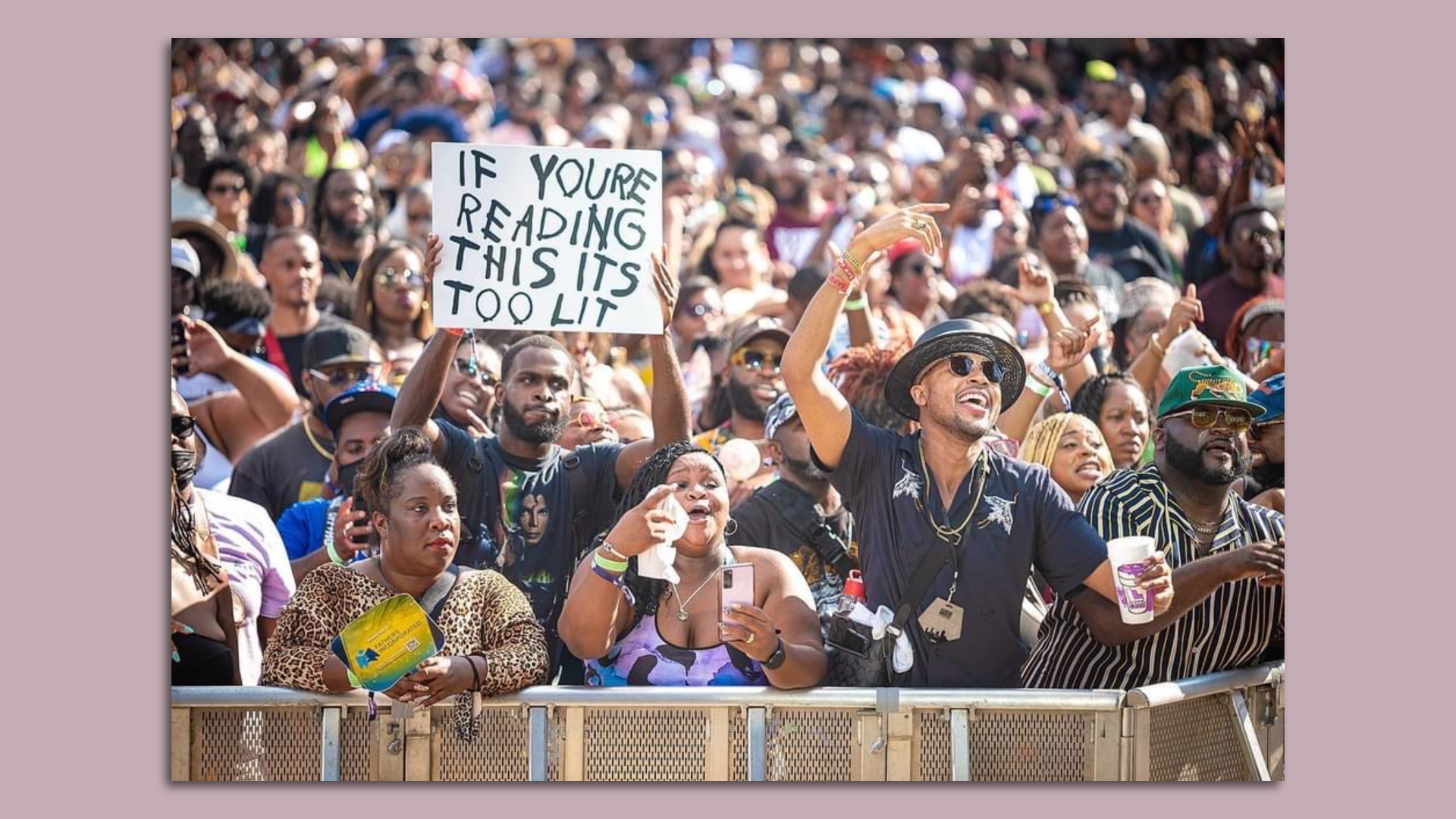 A large crowd of people enjoy a concert while a man holds a sign saying "if you are reading this it's too lit."