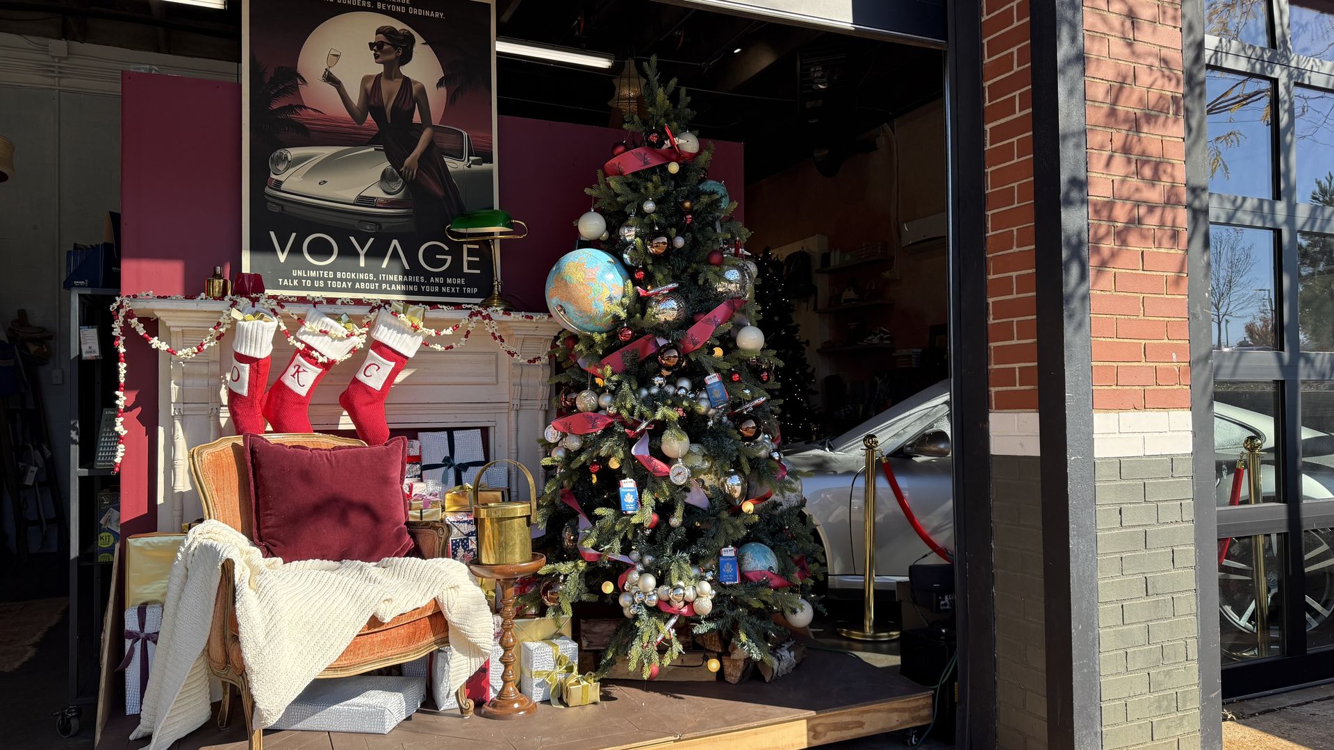 Christmas display with decorated tree featuring globes, ornaments, and red ribbons beside a chair draped with a cream blanket and red pillow, three red stockings on a faux fireplace, and wrapped gifts.