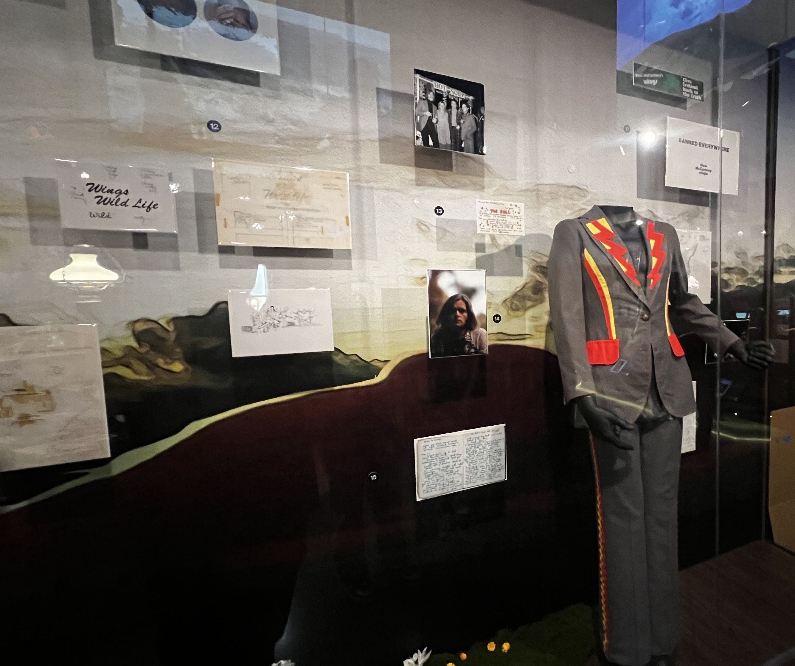 Museum display case with scattered documents and photos, a gray suit with red-and-yellow trim on a mannequin, and a black-and-white group photo; labels show 12–15.