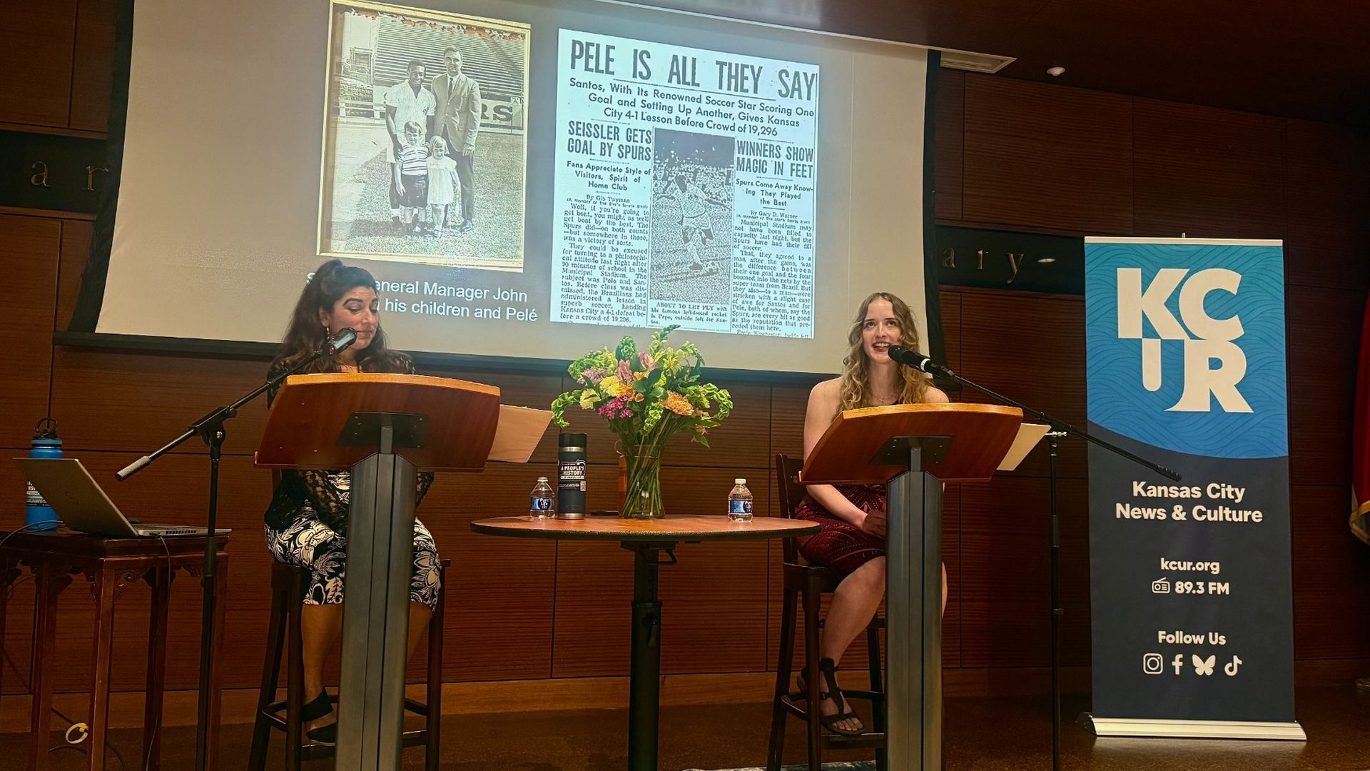 Two women seated at podiums on a stage with microphones, a projector screen displaying newspaper clippings, and a KCUR banner reading Kansas City News & Culture beside a flag; flowers on a table.
