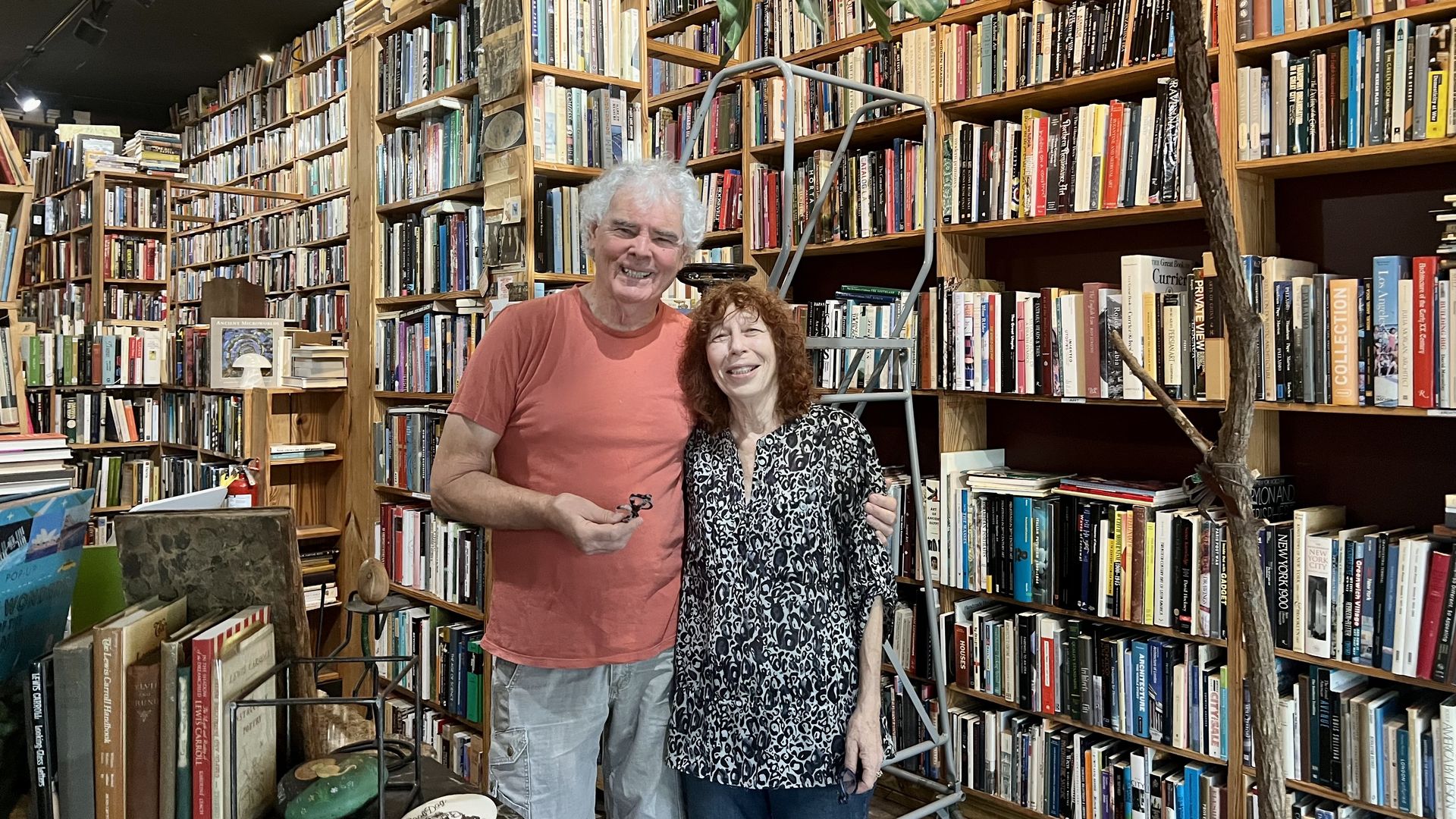An older man in a coral T-shirt and gray shorts stands with a woman in a black-and-white patterned blouse amid tall wooden bookshelves filled with books in a cozy bookstore.