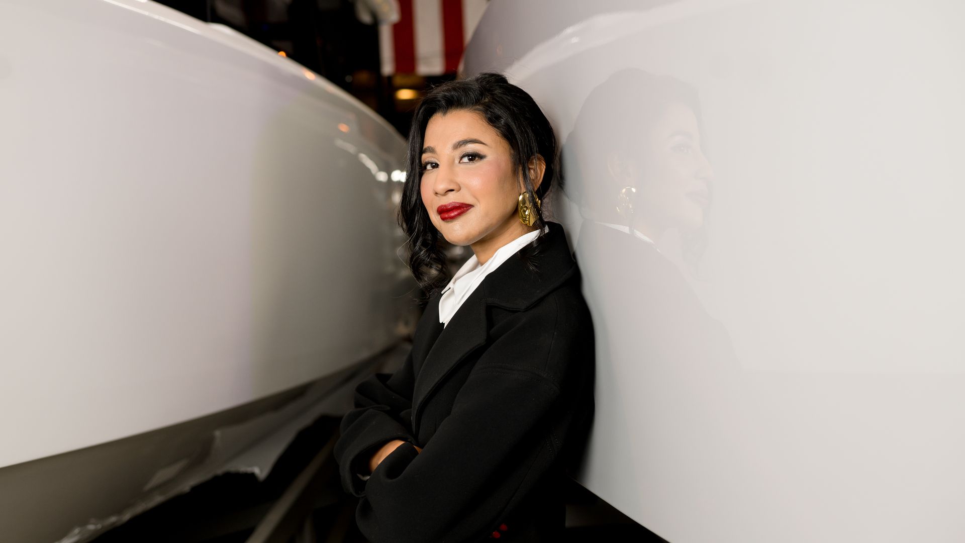 Woman with dark curly hair, red lipstick, and gold earrings wearing a black coat and white shirt, leaning against a glossy white surface with her arms crossed.
