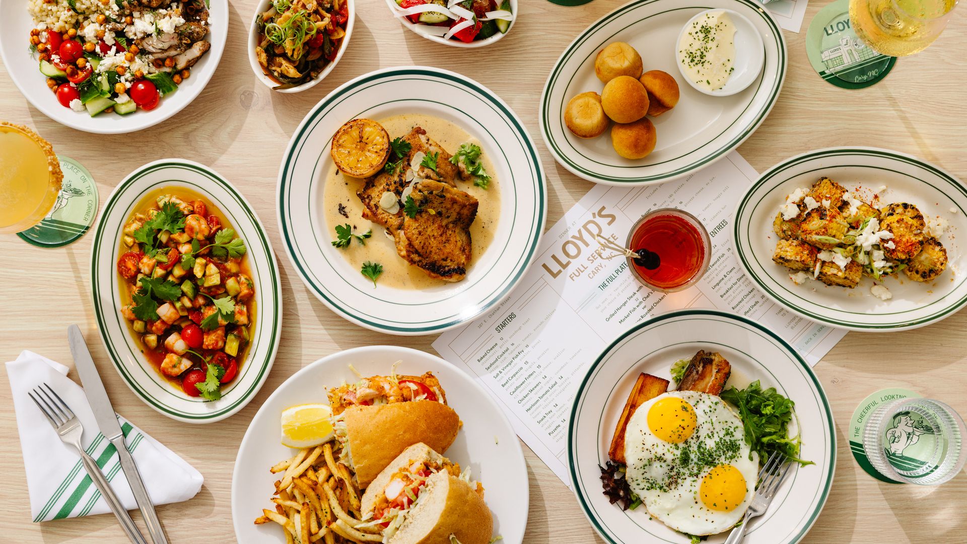Overhead view of a table loaded with green-rimmed plates: salads, roasted chicken with cream sauce, sautéed vegetables, breaded bites, eggs sunny-side up, fries, and drinks on coasters.