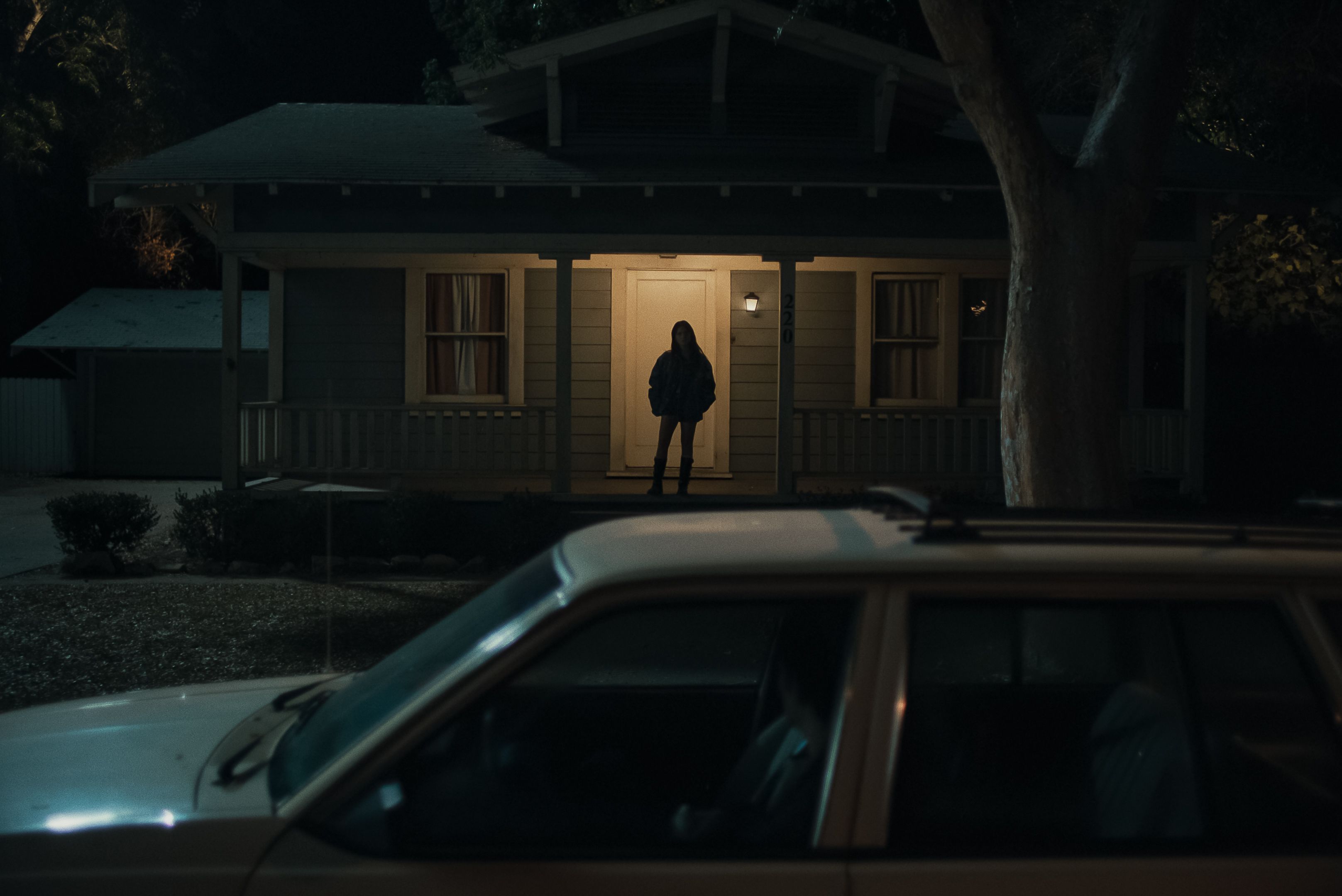 Nighttime scene of a small house with a lit front door. A silhouetted person in a jacket stands on the porch; a large tree to the right and a car hood in the foreground frame the shot.