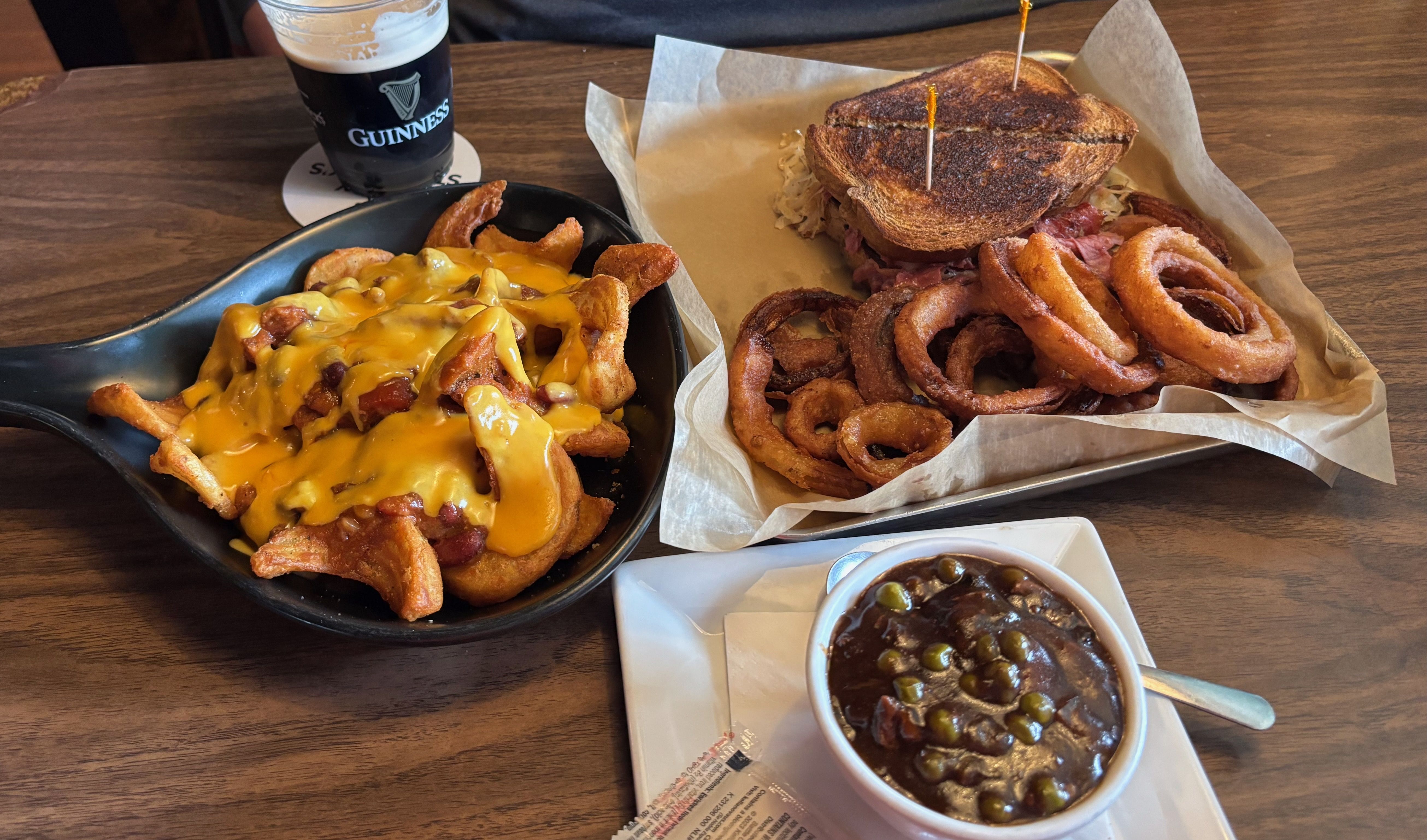 A wooden table with a skillet of cheese-covered loaded fries, a toasted Reuben sandwich with onion rings on the side, a cup of beef stew with peas, and a Guinness beer.
