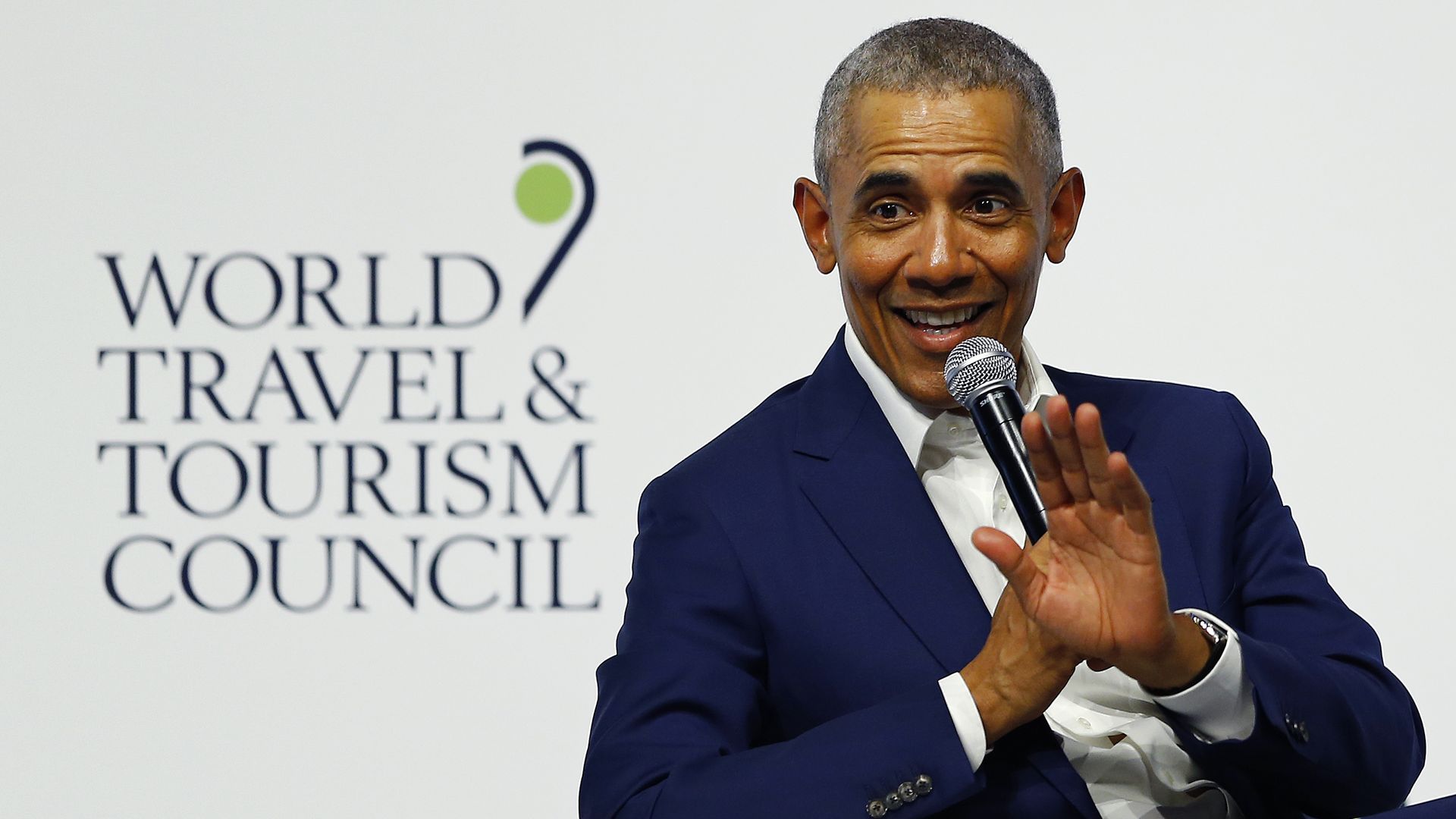 In this image, Obama sits in a chair and waves to the audience while smiling and speaking into a microphone.