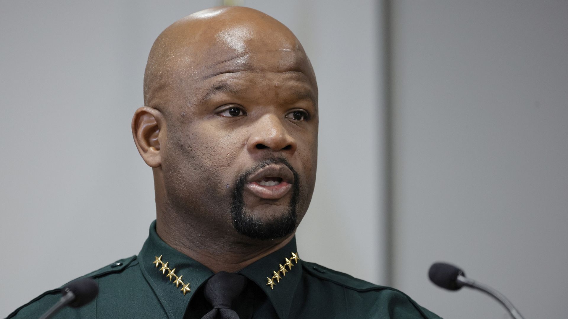 Sheriff Gregory Tony speaking behind two microphones wearing his green police uniform