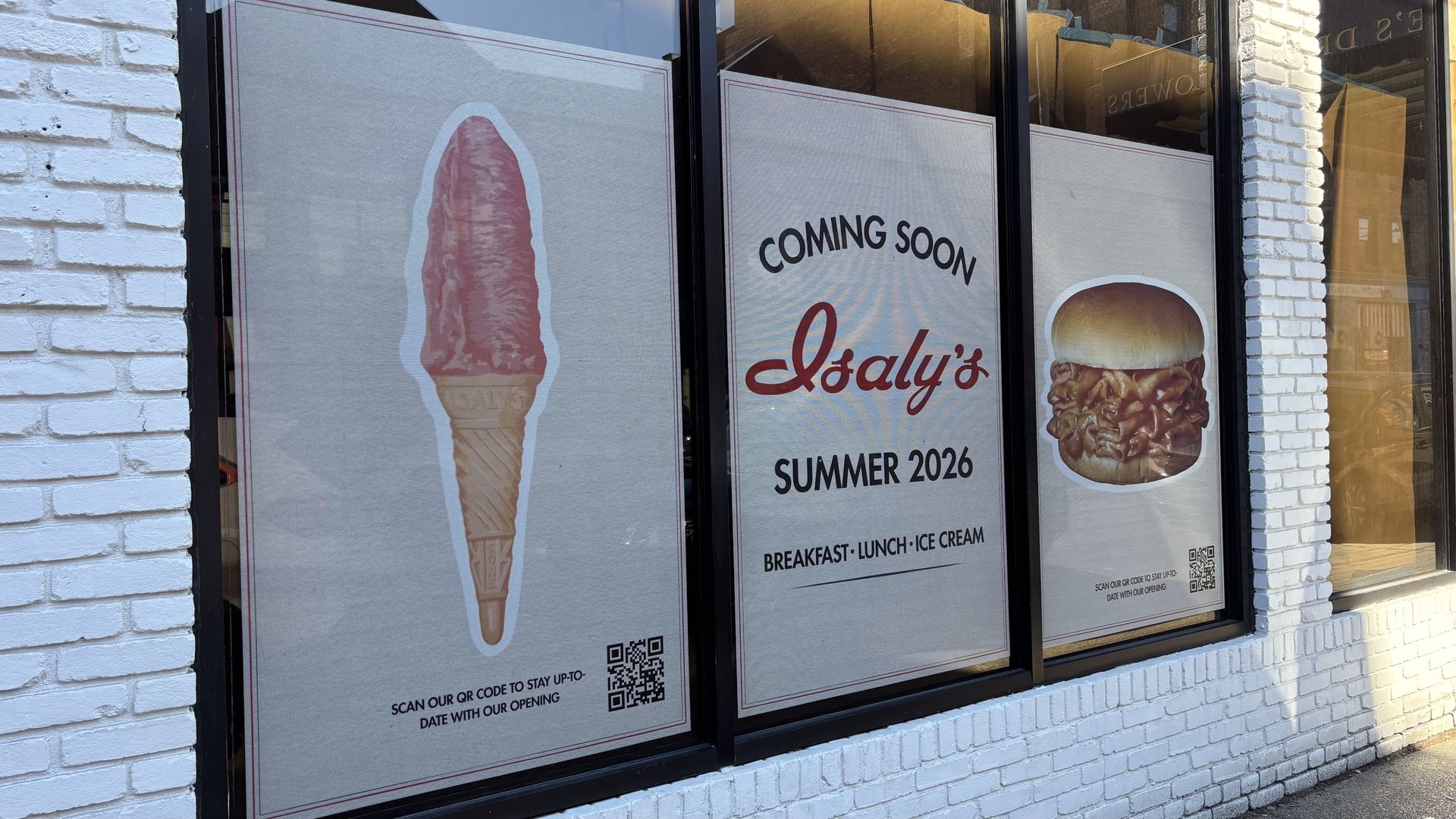 Window signs on a white brick wall announcing Isaly's coming summer 2026, showing a red ice cream cone and a sandwich with meat, offering breakfast, lunch, and ice cream.
