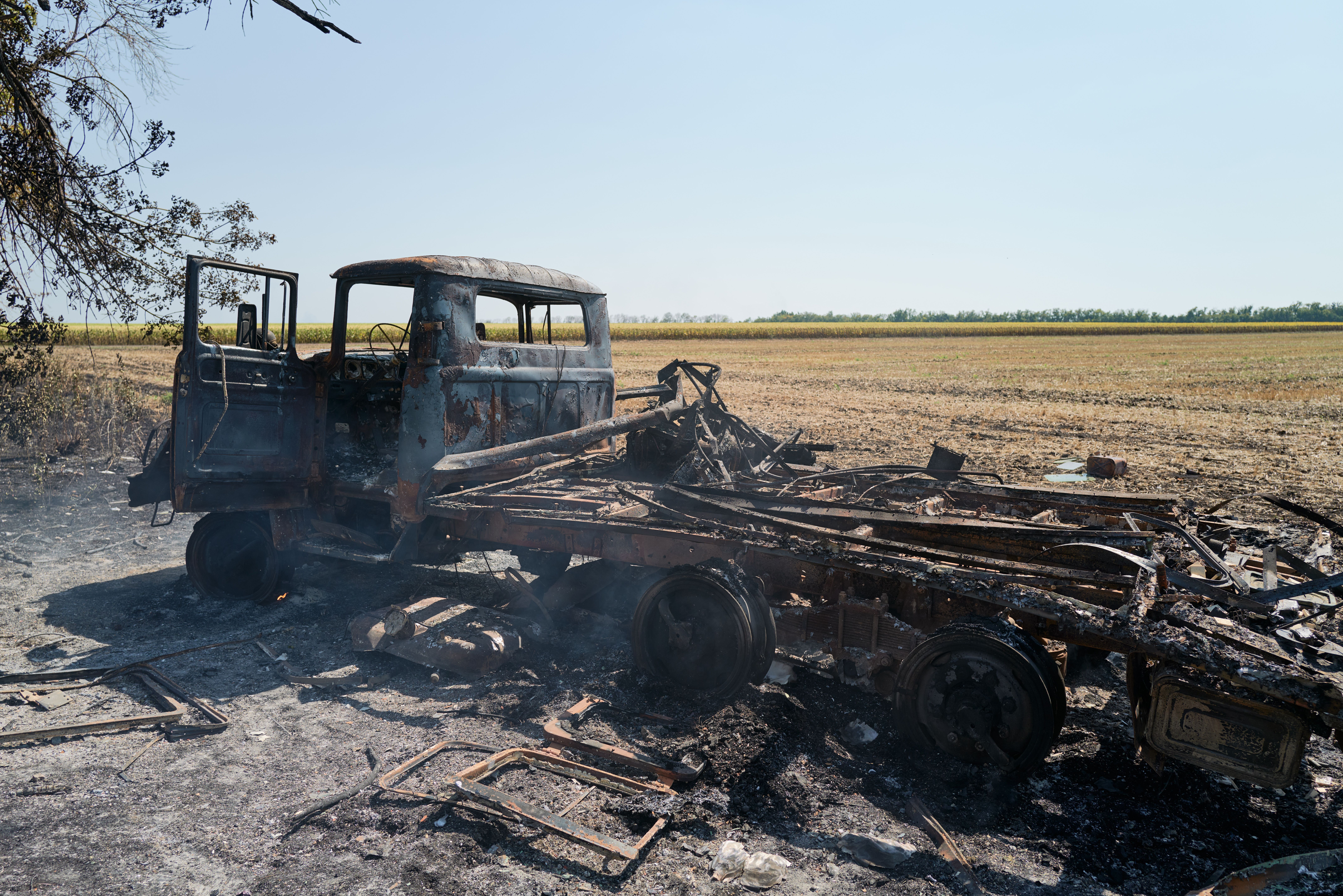 Burnt remains of a large truck with open door in a dry field under a clear blue sky, showing charred metal frame and wheelless axles, surrounded by ashes and debris.