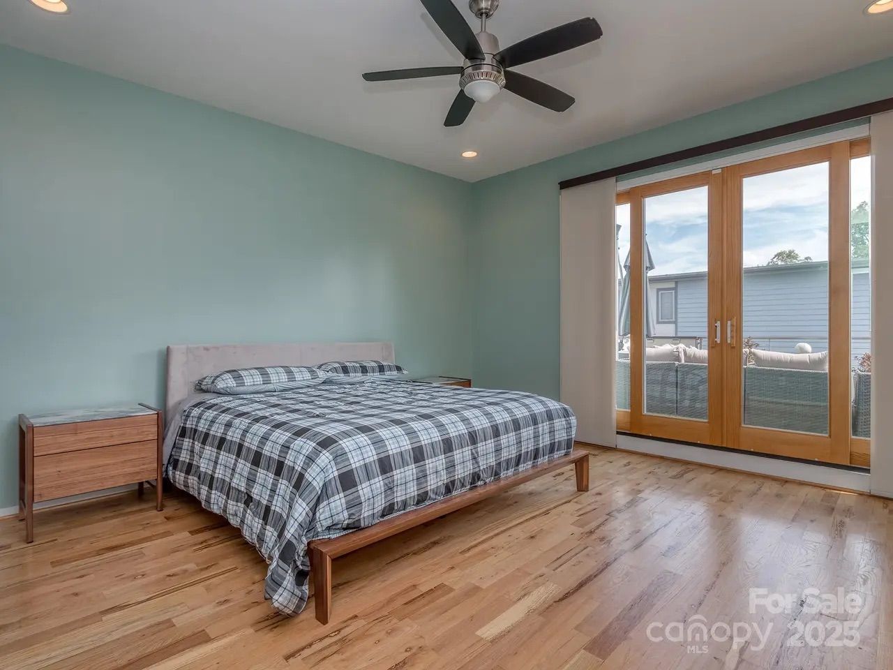 Bedroom with light blue walls, wooden floor, king-size bed with black-and-white plaid bedding, two wooden nightstands, ceiling fan, and large wooden framed glass doors leading outside.