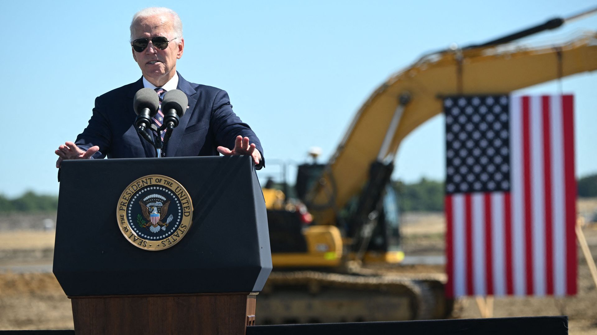President Biden speaks at the Intel groundbreaking in front of a tractor draping an American flag.