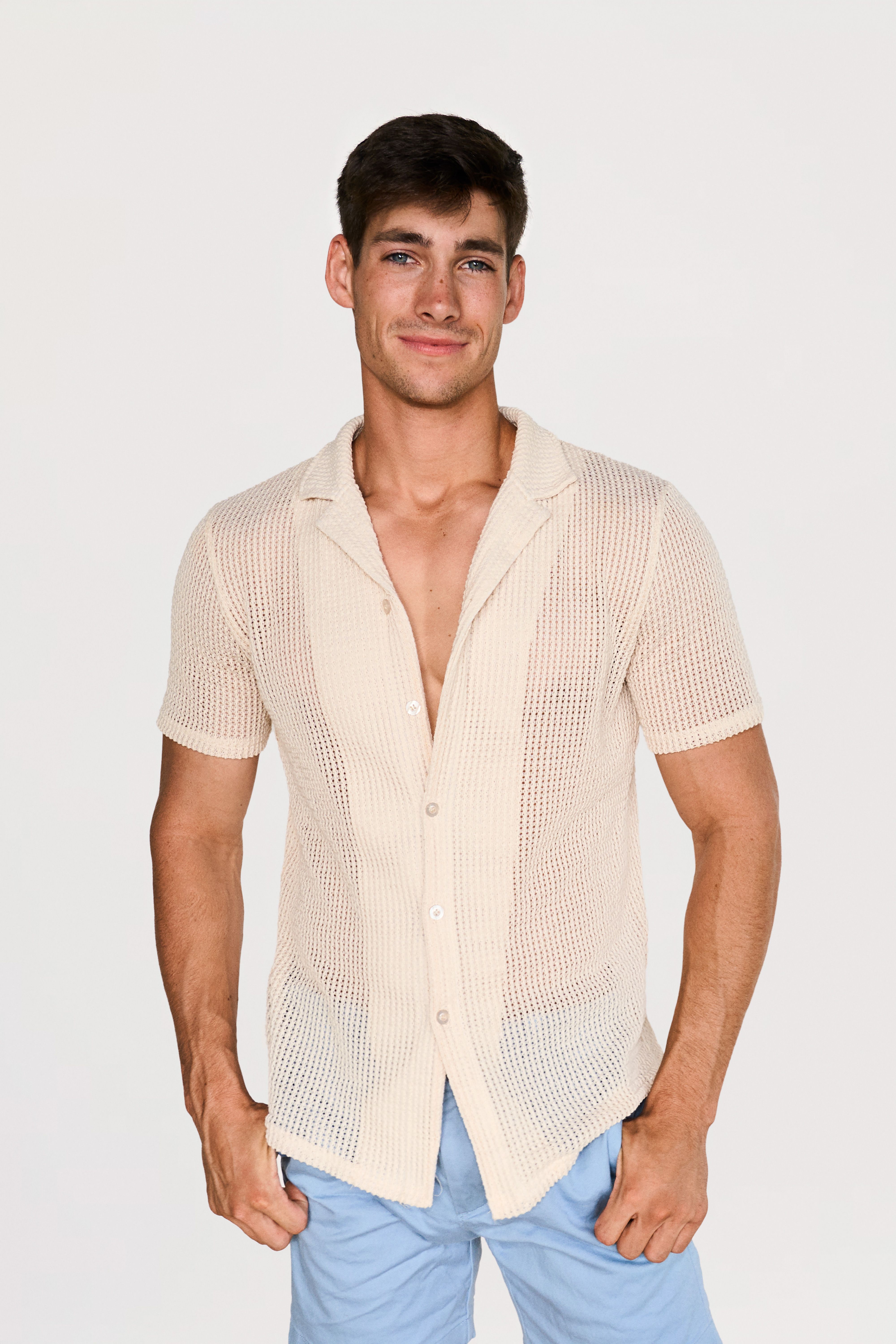 Young man with dark hair wearing a beige open-knit short-sleeve shirt and light blue shorts, standing against a plain white background, smiling slightly.