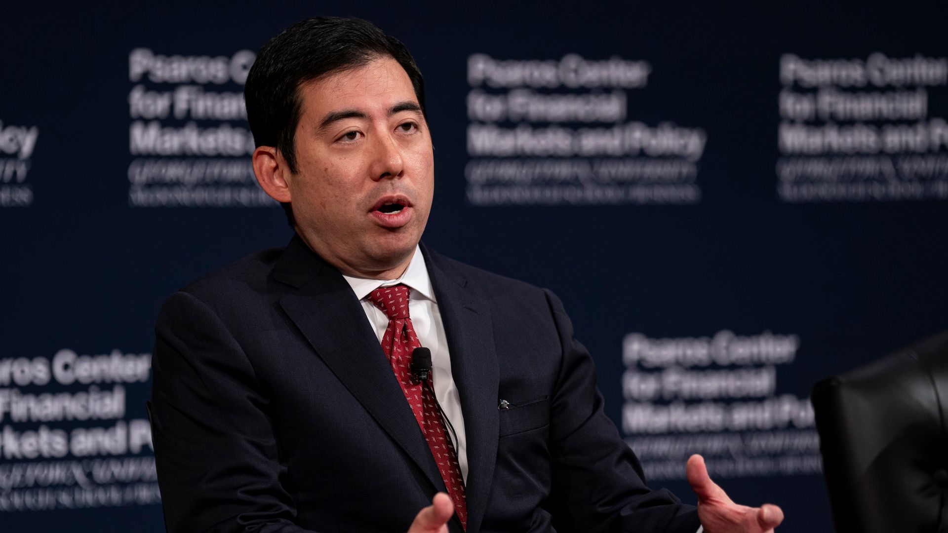 A man speaking at a conference, sitting down, with a red tie.
