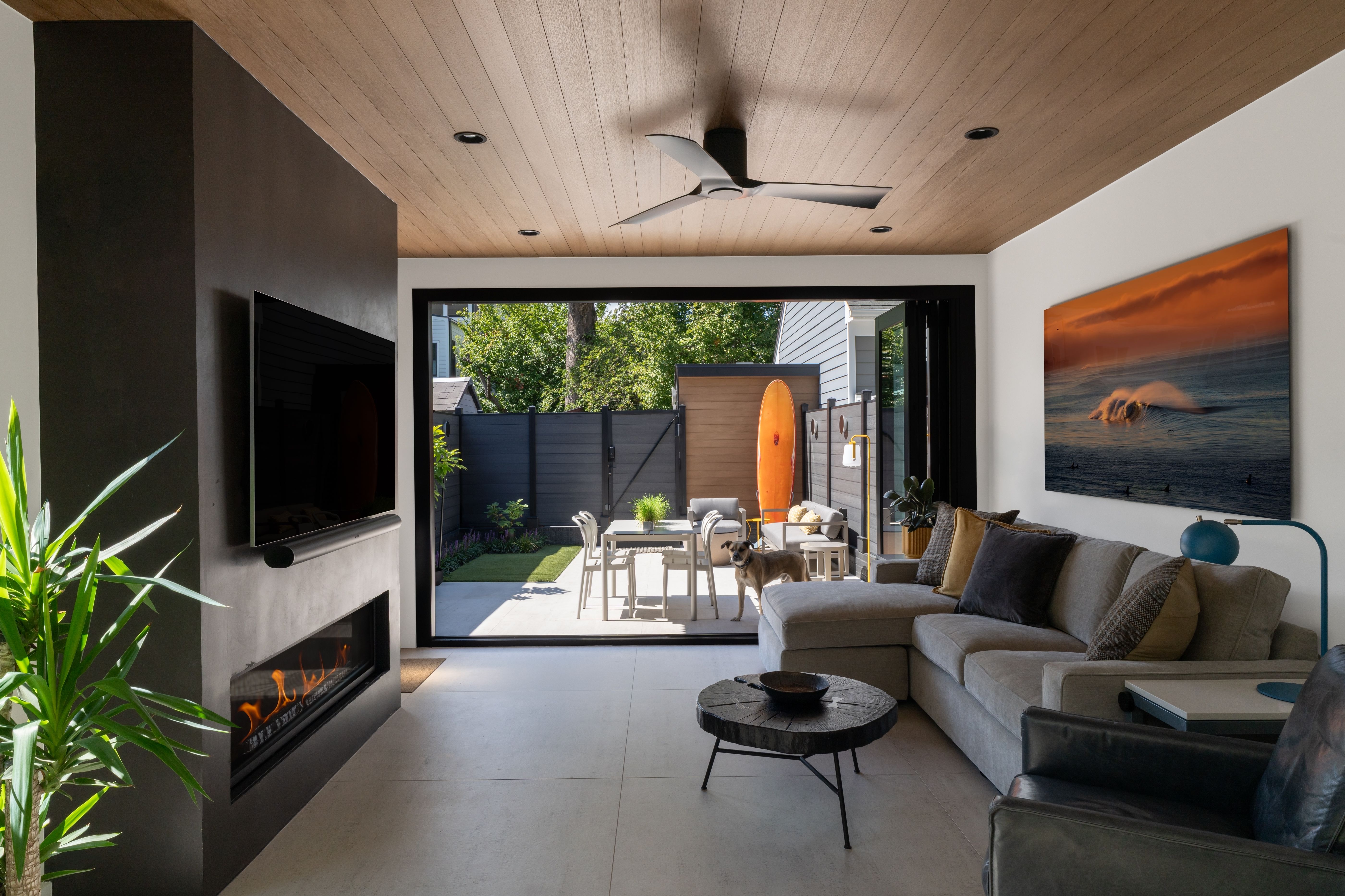Modern living room with a wood ceiling, black wall, wall-mounted TV, and linear fireplace. A gray sectional faces the TV; sliding doors open to a patio with a surfboard, a dog, and outdoor seating.