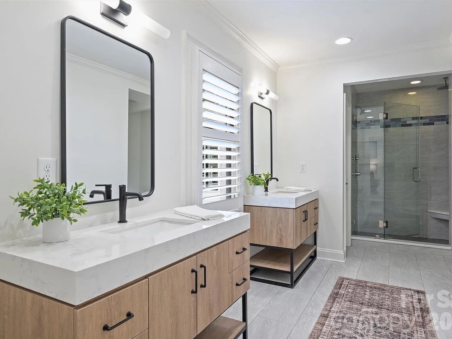 Modern bathroom with dual wooden vanities, white marble countertops, black fixtures, rectangular mirrors, a glass shower with gray tiles, white walls, and a brown rug on light gray floor tiles.