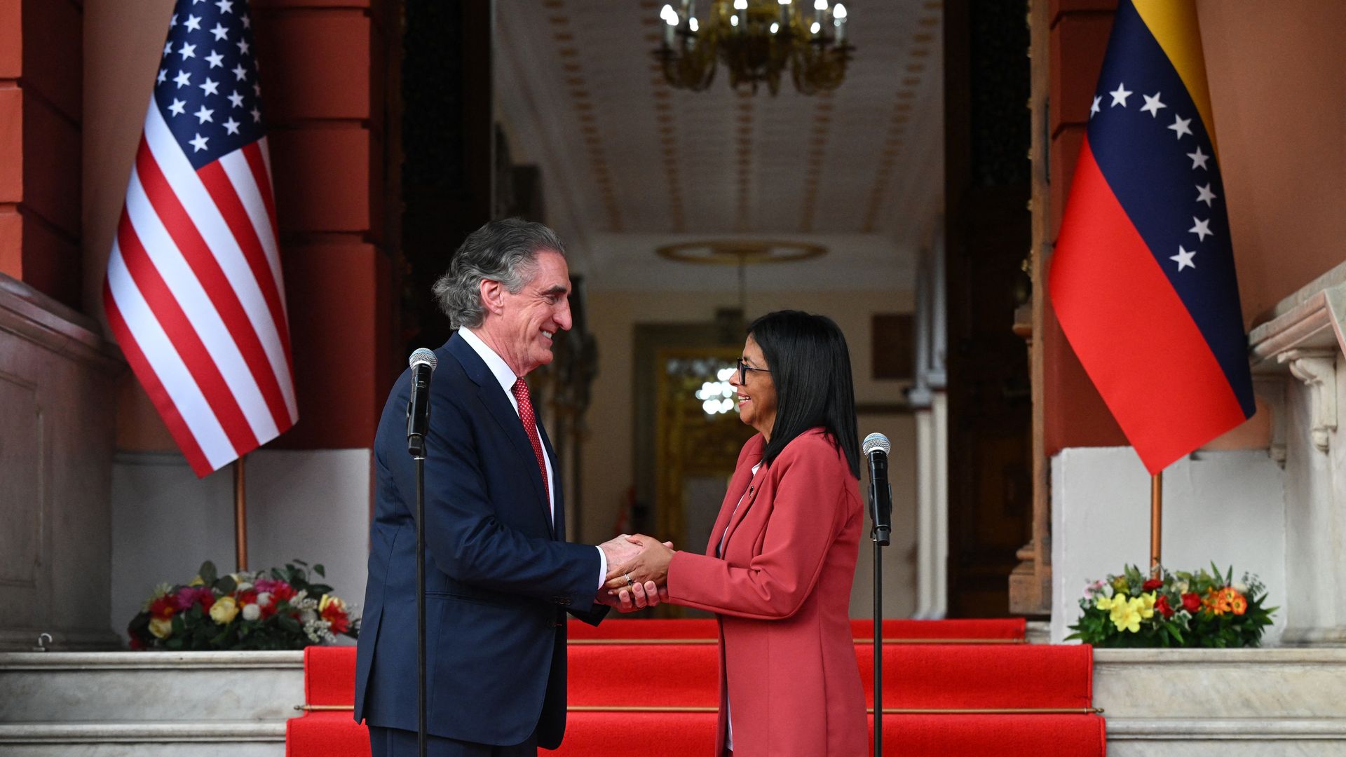 Interior Secretary Doug Burgum and Venezuela's interim president, Delcy Rodriguez, in formal attire shaking hands on red carpeted stairs between U.S. and Venezuelan flags with flower arrangements, microphones in front, inside a grand building.