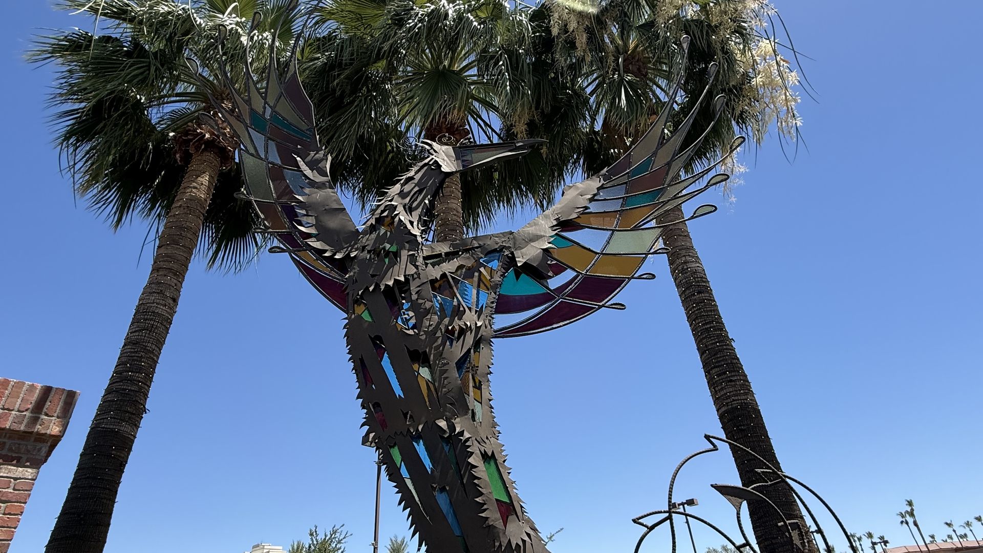 Metal sculpture of a bird with outstretched wings made from multicolored glass panels; two palm trees rise behind the bird to its left and right.