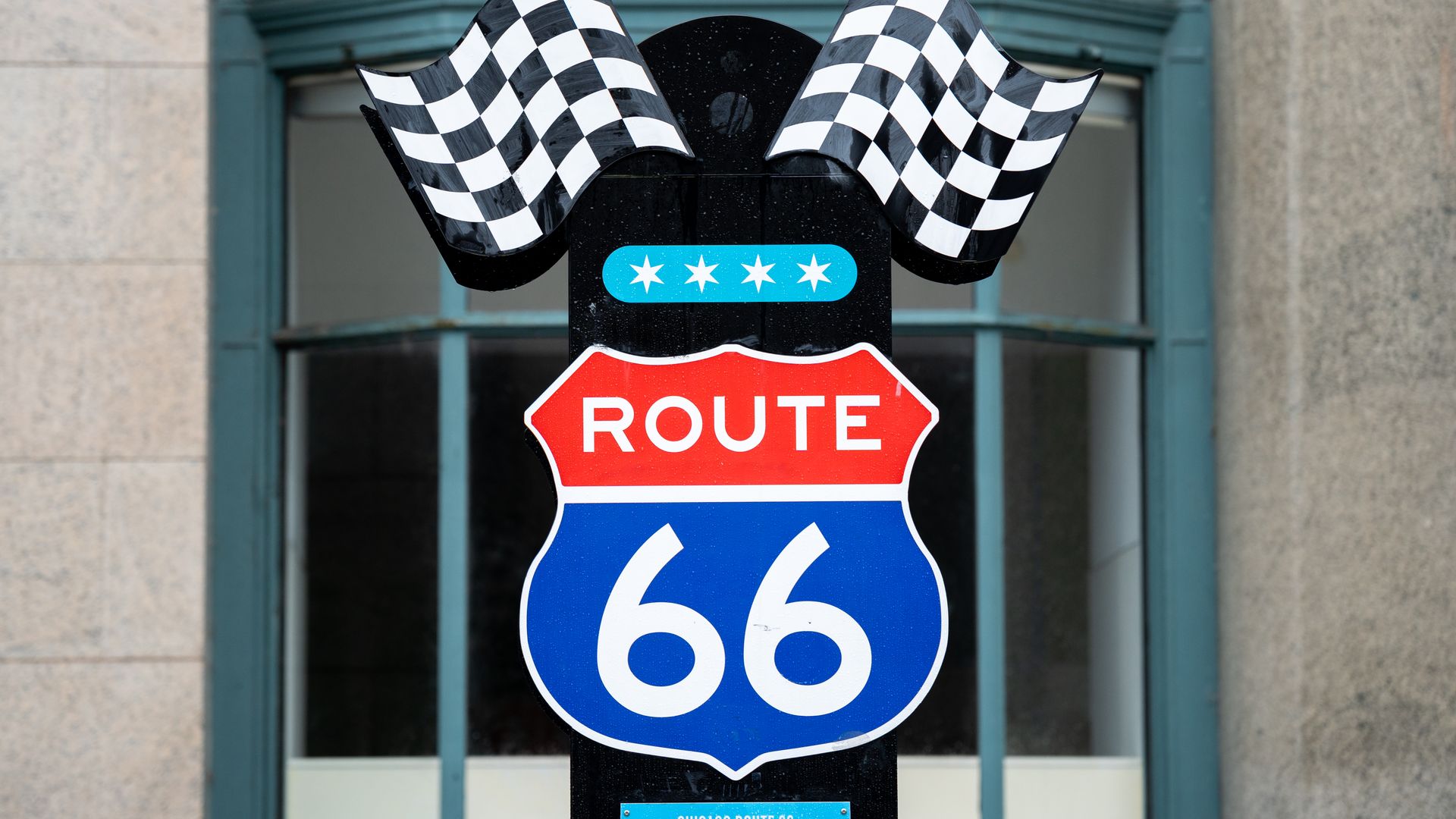 Close-up of a Route 66 signpost topped with black-and-white checkered flags, displaying a red 'ROUTE' shield and a blue '66' shield, with a small blue trail plaque below, set against a stone building.