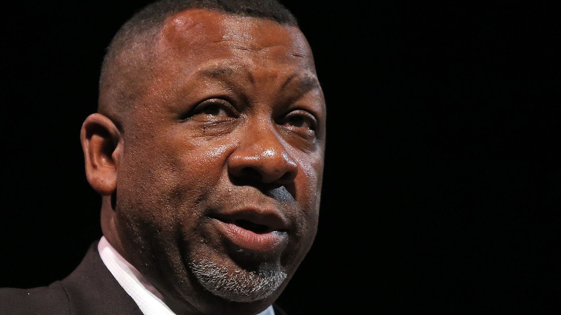 Close-up of a middle-aged Black man wearing a dark suit, white shirt, and patterned tie, speaking against a black background.