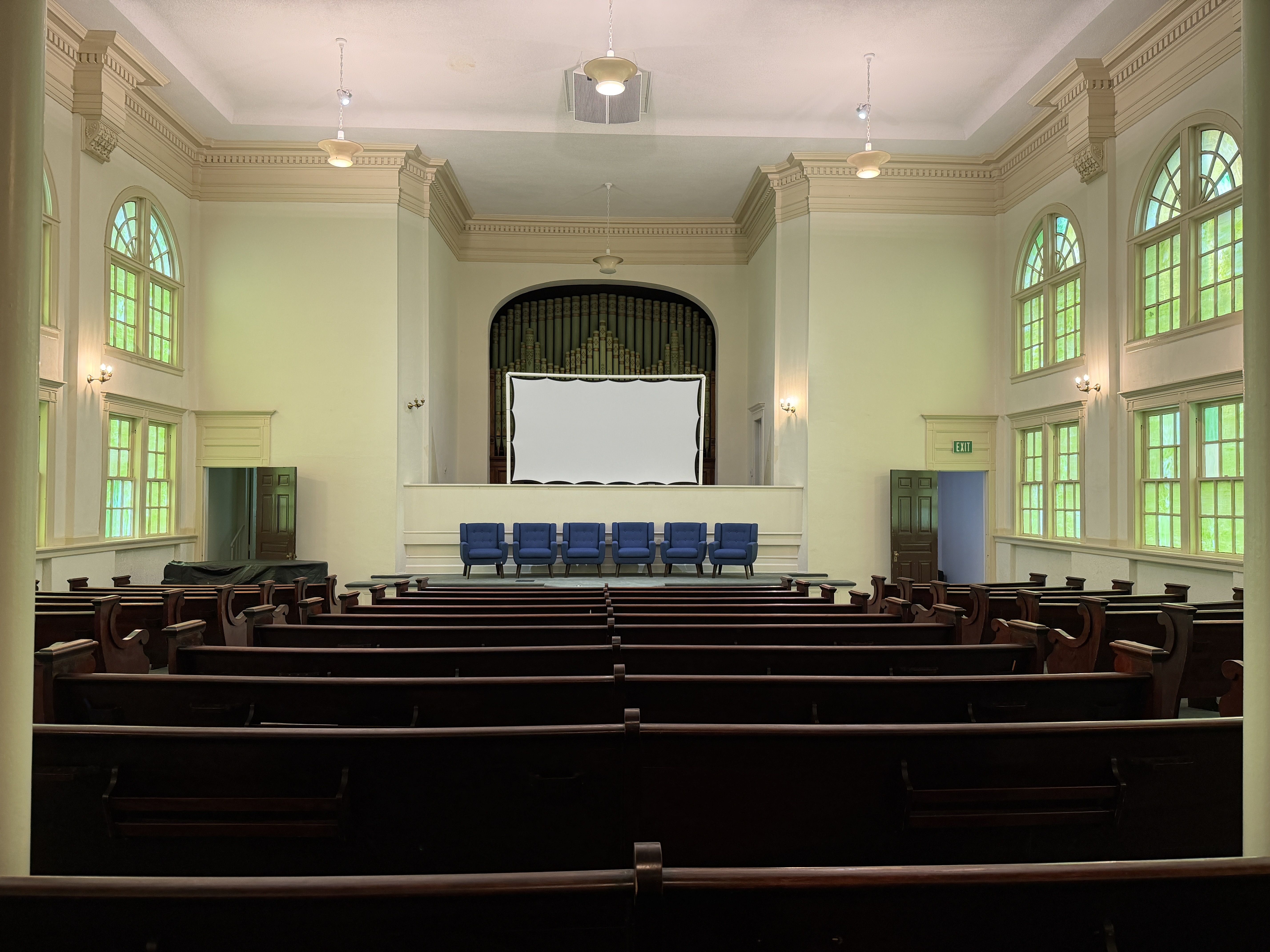 Interior of a sunlit, classical hall with arched windows and cream molding. A raised stage features a pipe organ behind a white screen, with blue chairs and dark wooden pews.