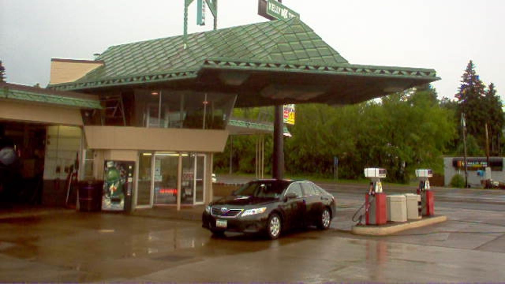 Gas station with a green roof and two red fuel pumps, a black car parked nearby, and a sign reading "SPUR" and "Kelly Rd" on a cloudy day.
